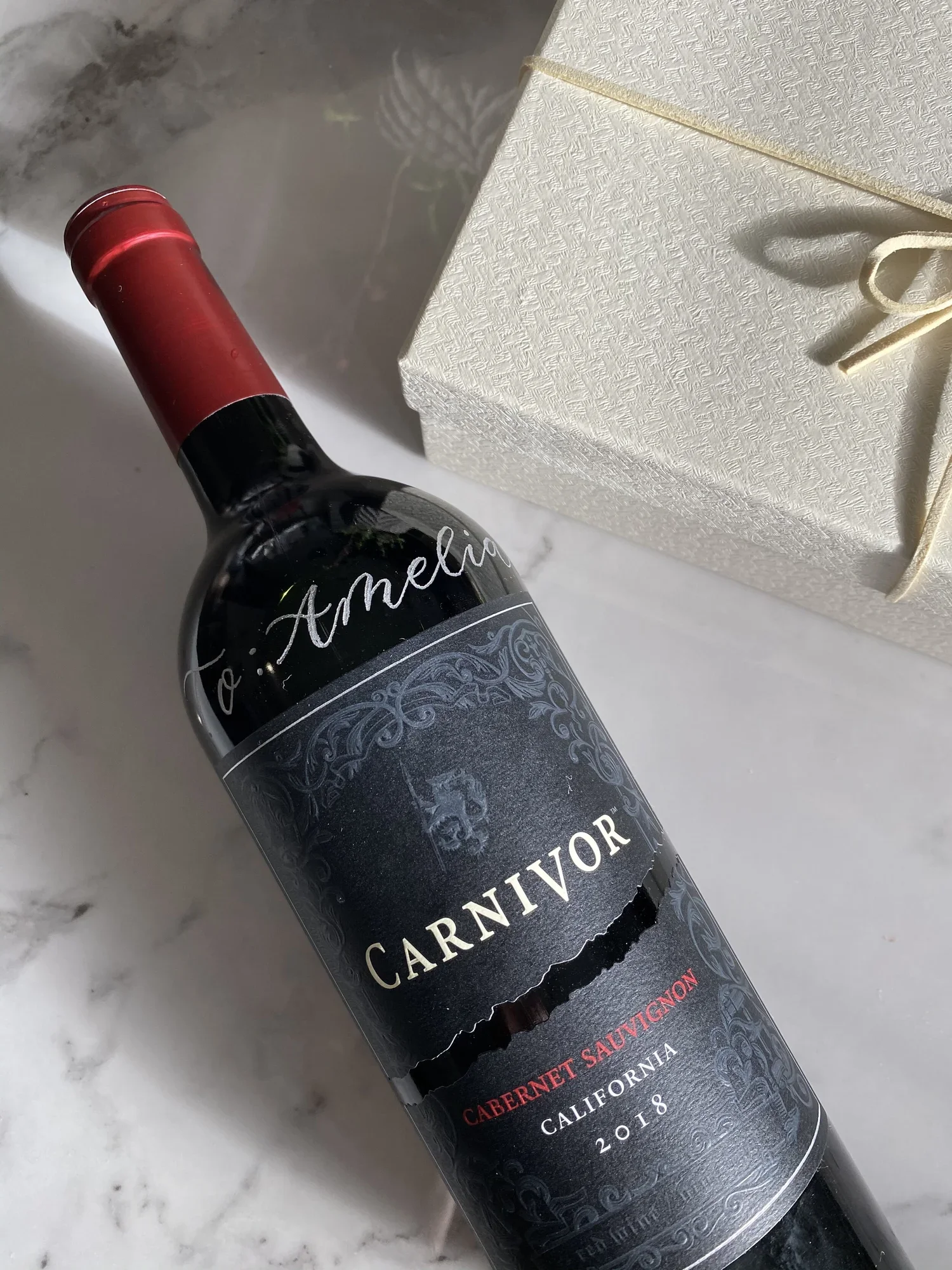 A bottle of Carnivor Cabernet Sauvignon red wine from California, vintage 2018, placed on a marble surface next to a cream-colored gift box.