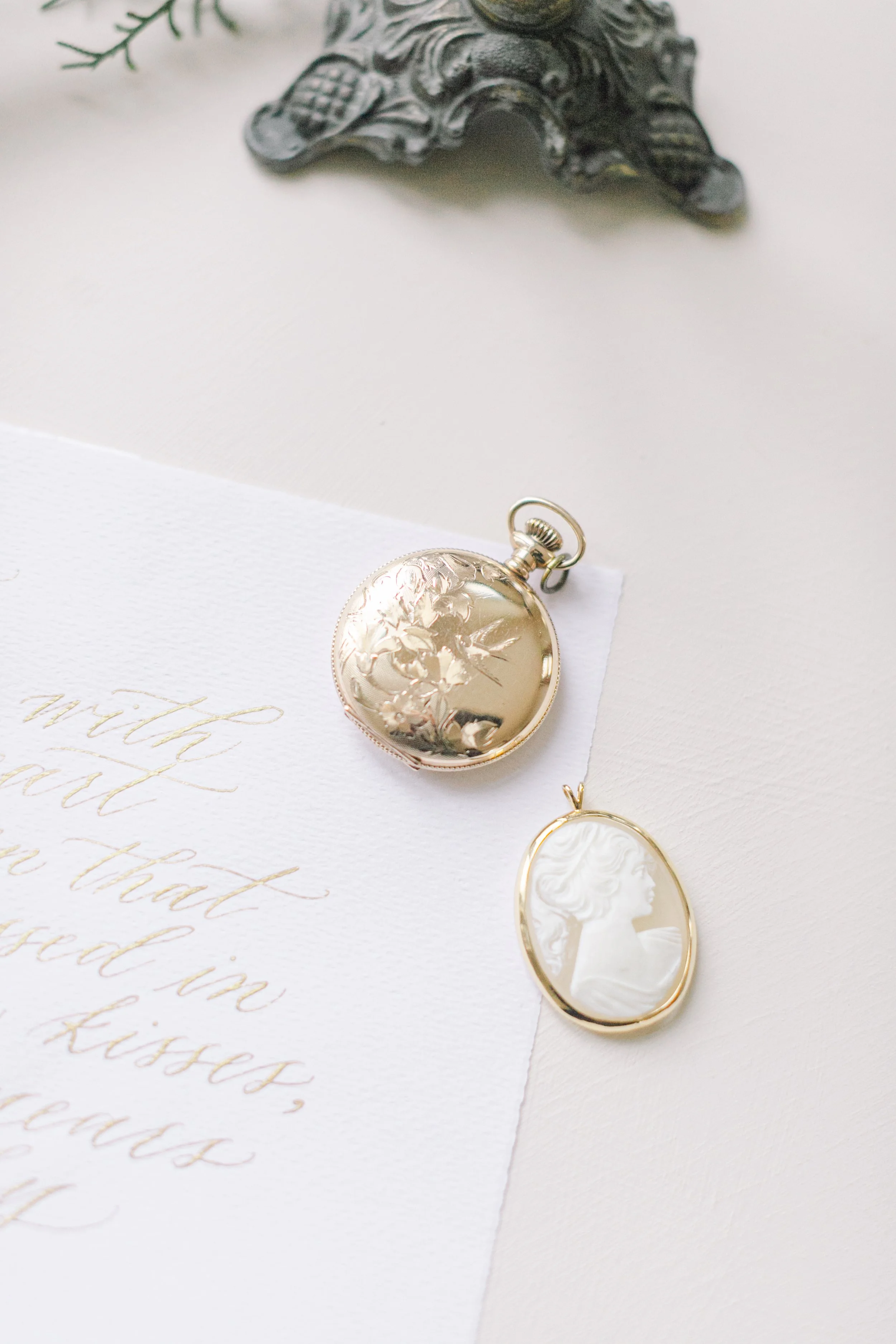 A vintage gold pocket watch and a cameo brooch on a white textured paper with elegant gold script, a decorative metal ornament, and a sprig of greenery in the background.