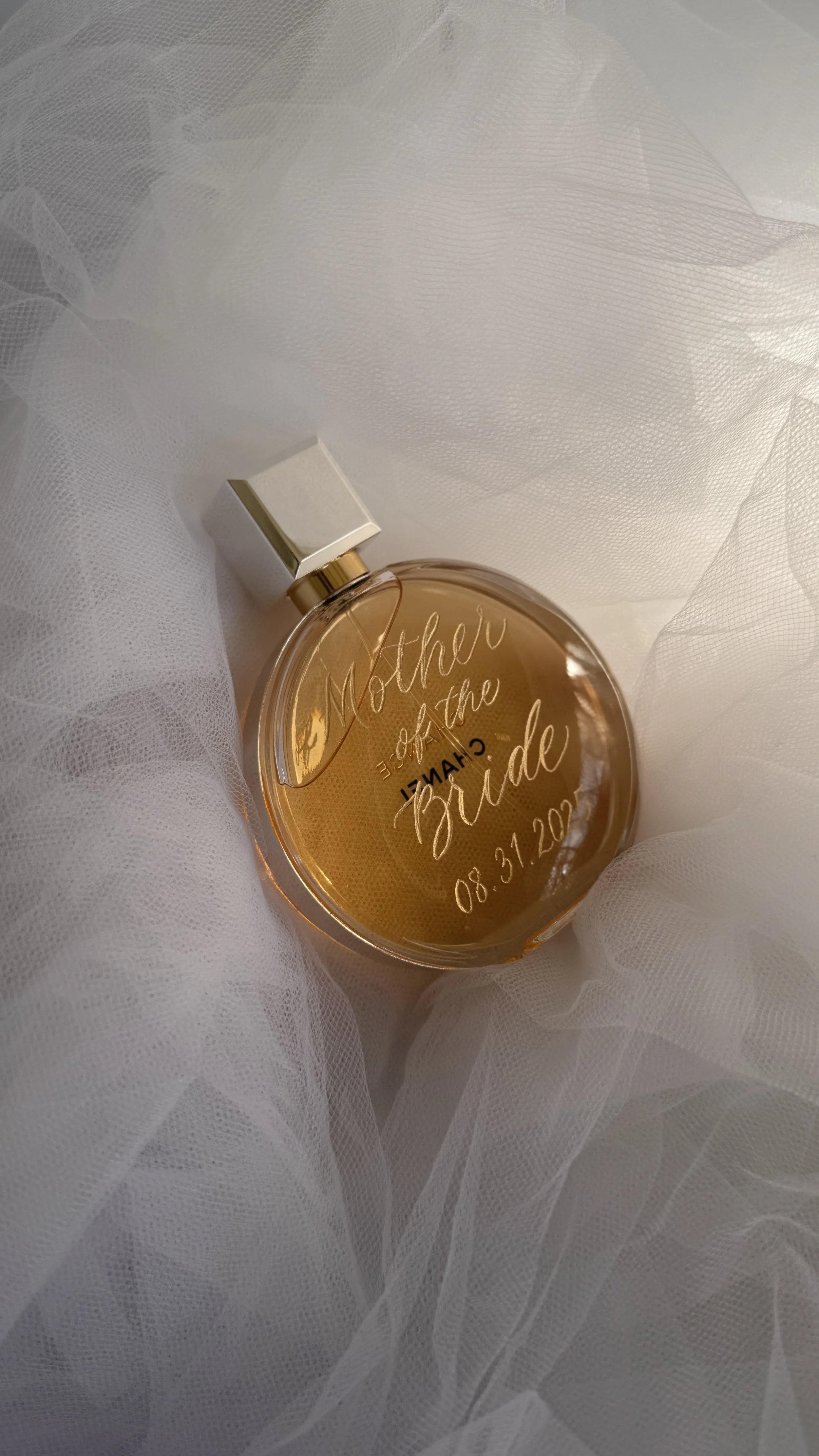 A round perfume bottle with a gold cap and orange liquid inside, resting on white tulle fabric. The bottle has engraved text that says, 'Mother of the Bride 08.31.2023'.