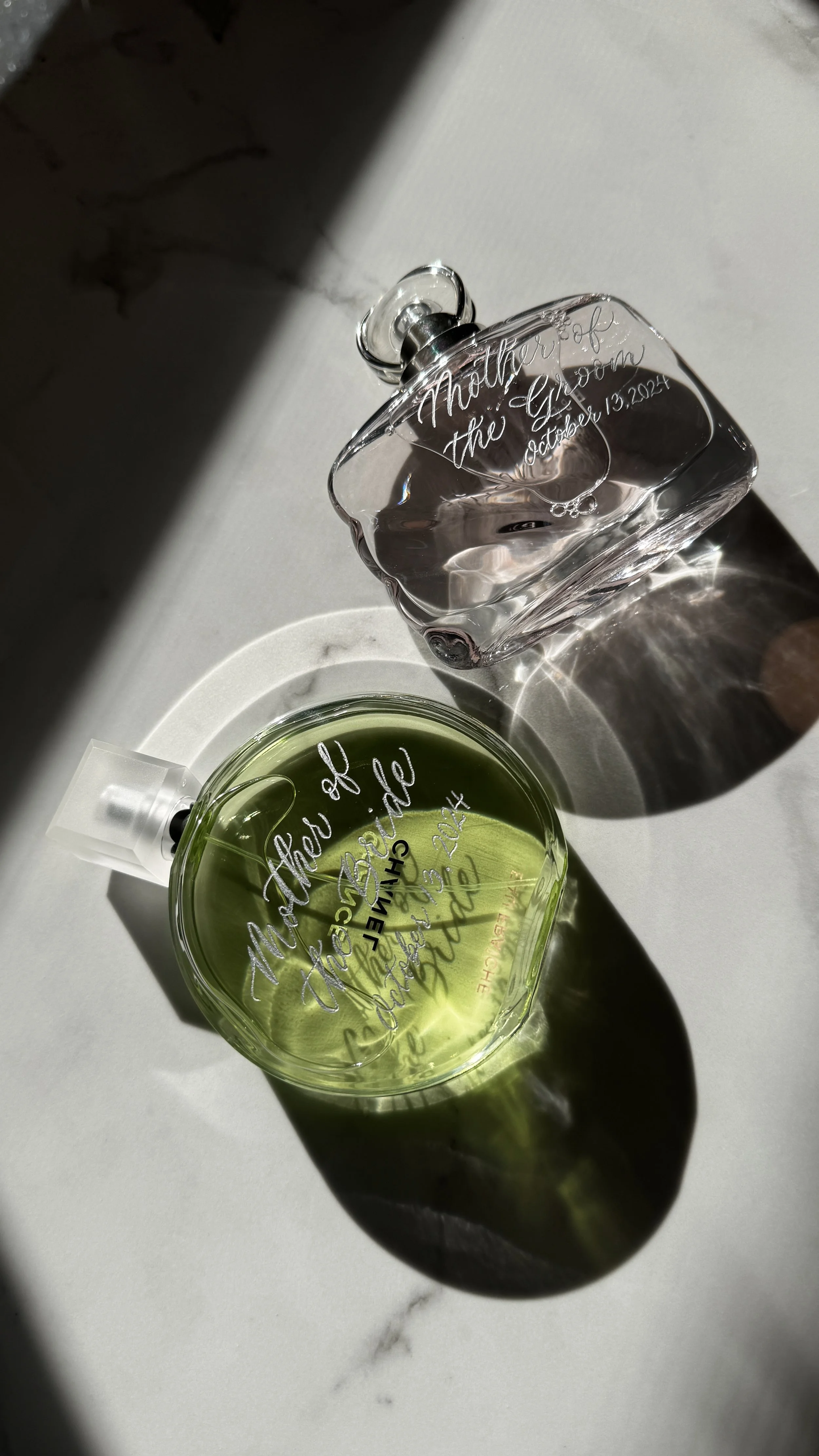 A perfume bottle and a glass perfume container on a white surface with engraved writing and a shadow cast from natural sunlight.