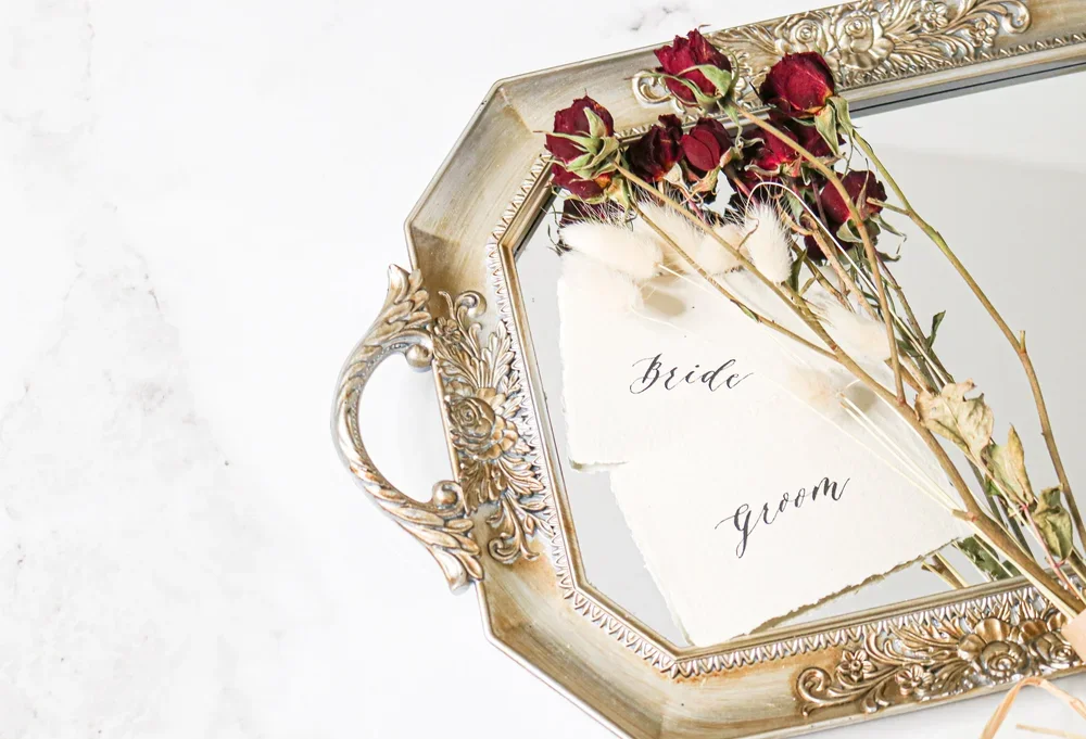 A decorative silver tray with ornate handles, holding torn paper with the words 'bride' and 'groom' written on it. Dried red roses are placed on top of the paper.