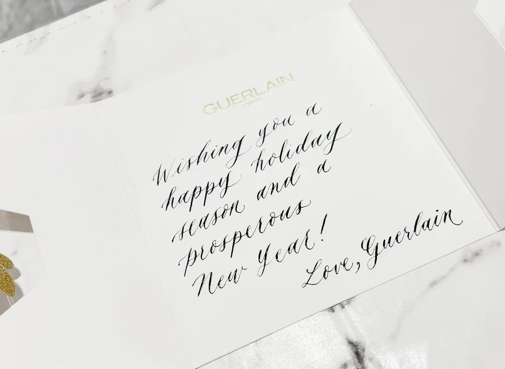 A holiday greeting card with the message: "Wishing you a happy holiday season and a prosperous New Year! Love, Guerlain" written in black cursive, gold embossed "GUERLAIN PARIS" at the top, on a marble surface.