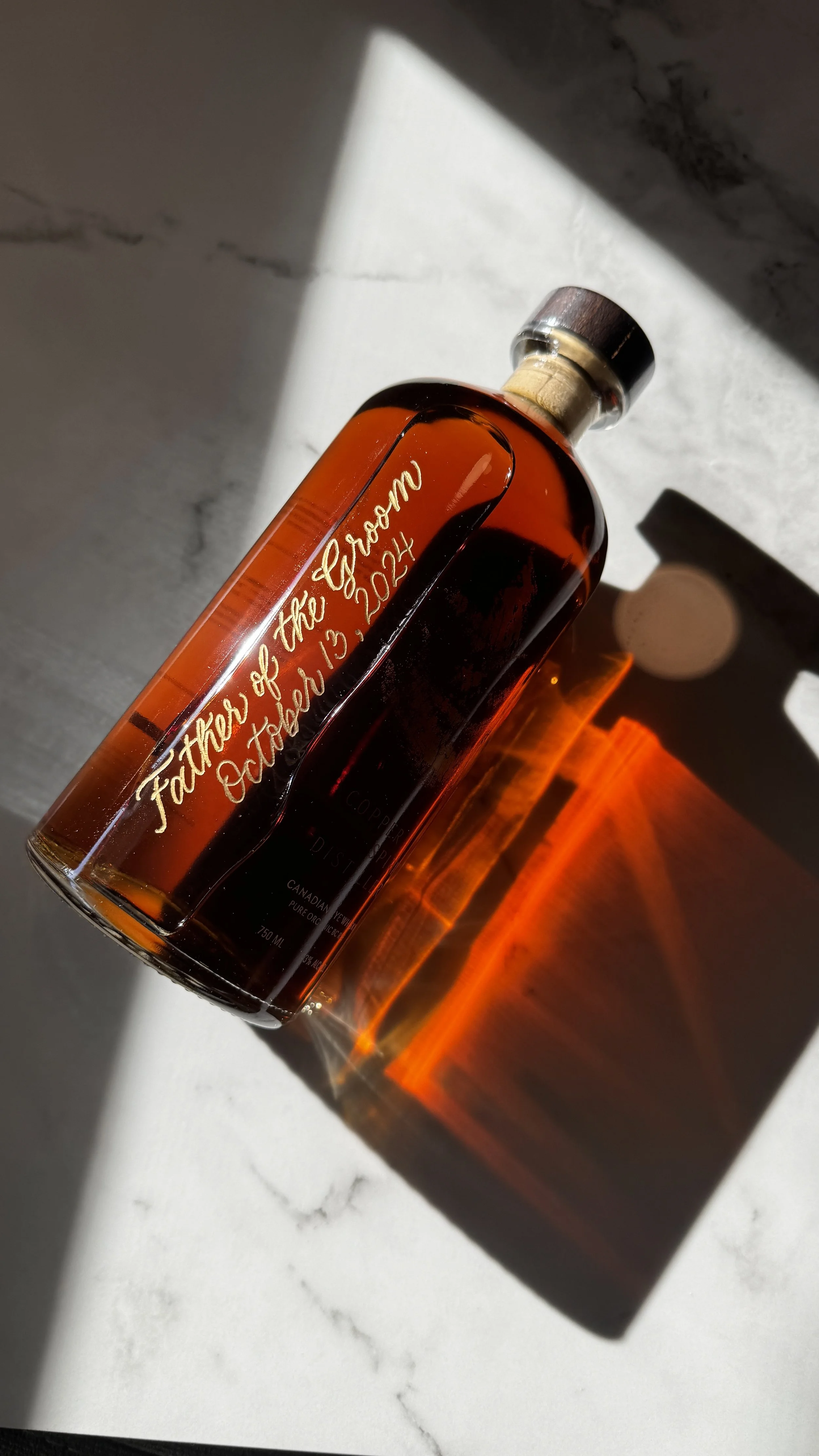 A glass bottle with orange liquid, illuminated by sunlight, with the inscription "Father of the Groom October 13, 2024" engraved on it, casting a shadow on a white surface.