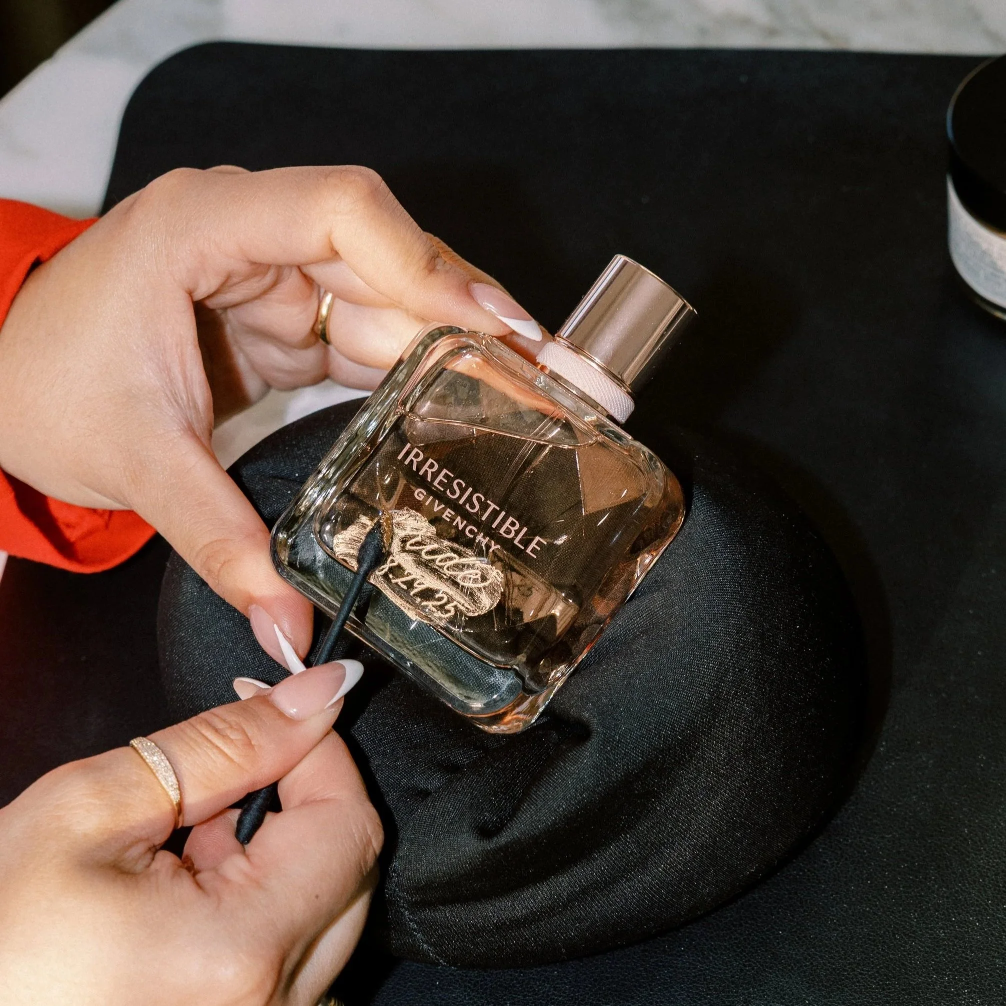 Person holding a bottle of Givenchy perfume labeled 'Irresistible' while removing the sprayer cap with their other hand. The person has manicured nails and is wearing a gold ring and a gold ring. The background includes a black surface and a small container.