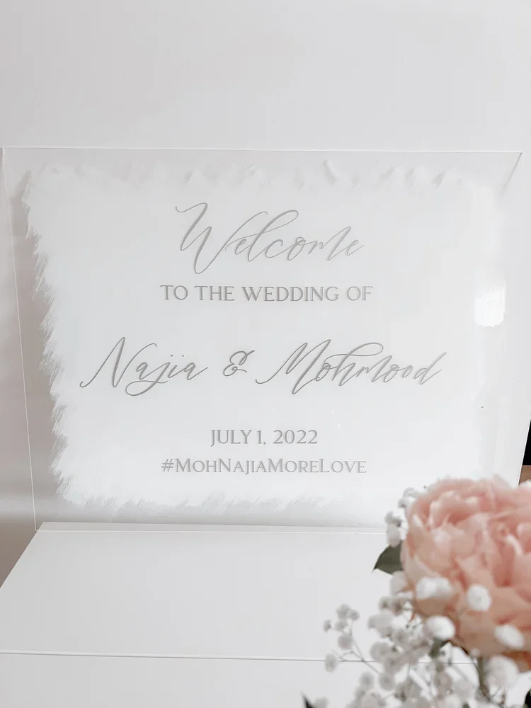 A clear wedding welcome sign with elegant script text, reading "Welcome to the wedding of Naija & Mothwood
July 1, 2022
#MohNajaMoreLove." The sign is placed on a white surface with pink and white flowers in the foreground.