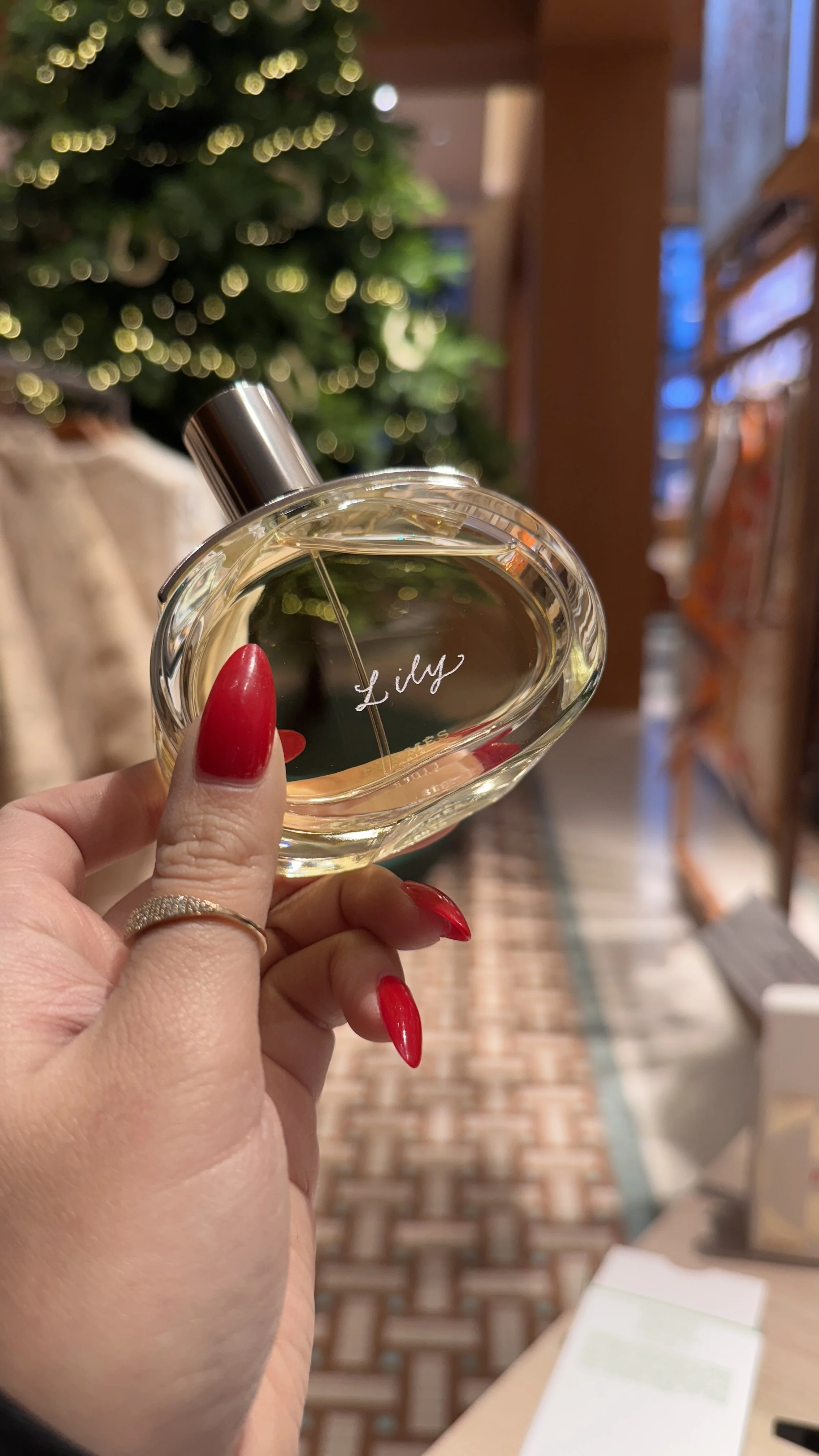 A hand with red painted nails holding a glass perfume bottle labeled 'Lidy' in front of a decorated Christmas tree with blurred lights in the background.