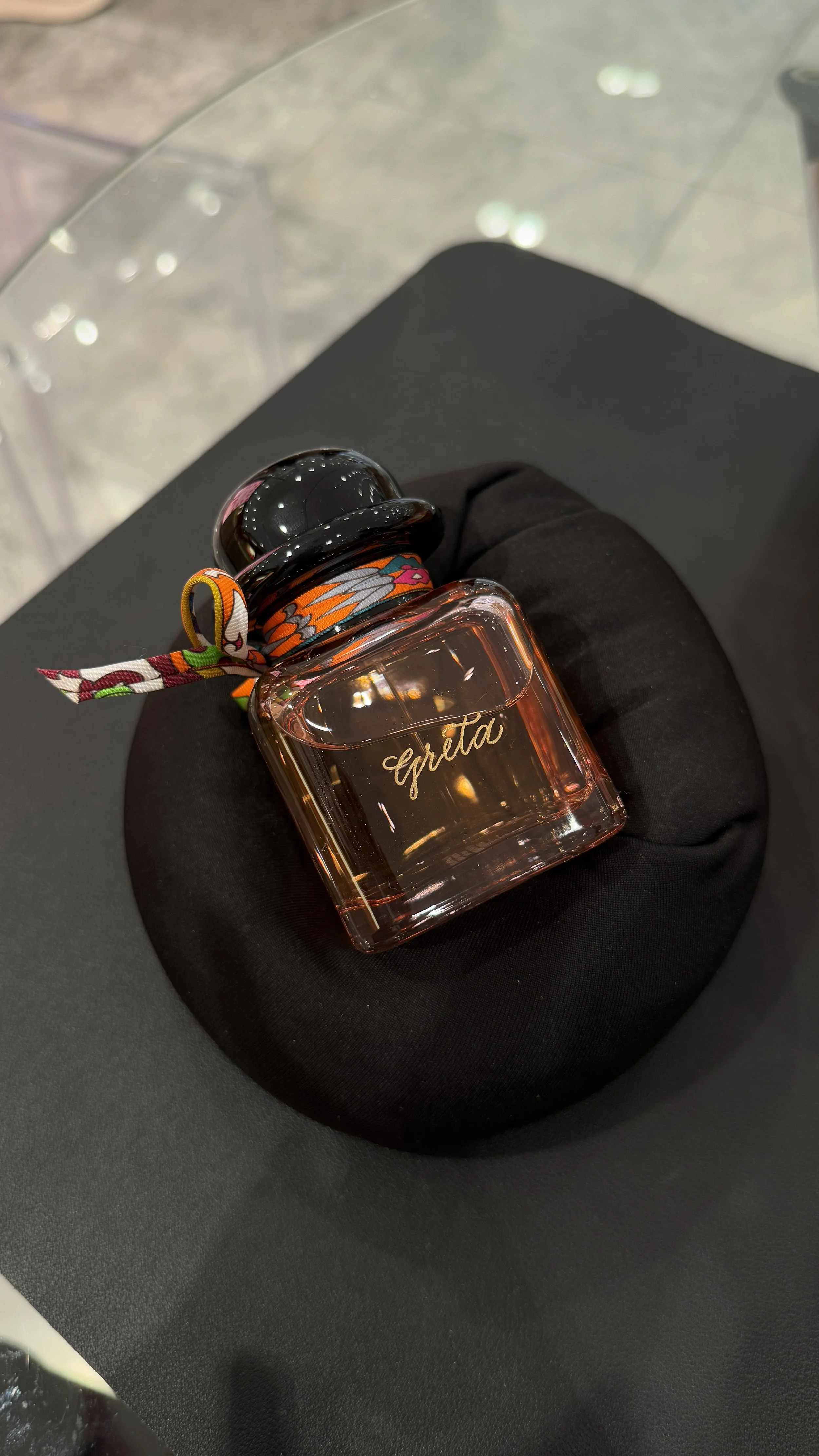 A rectangular glass perfume bottle with the word 'eleta' written on it, placed on a black cloth on a glass table.