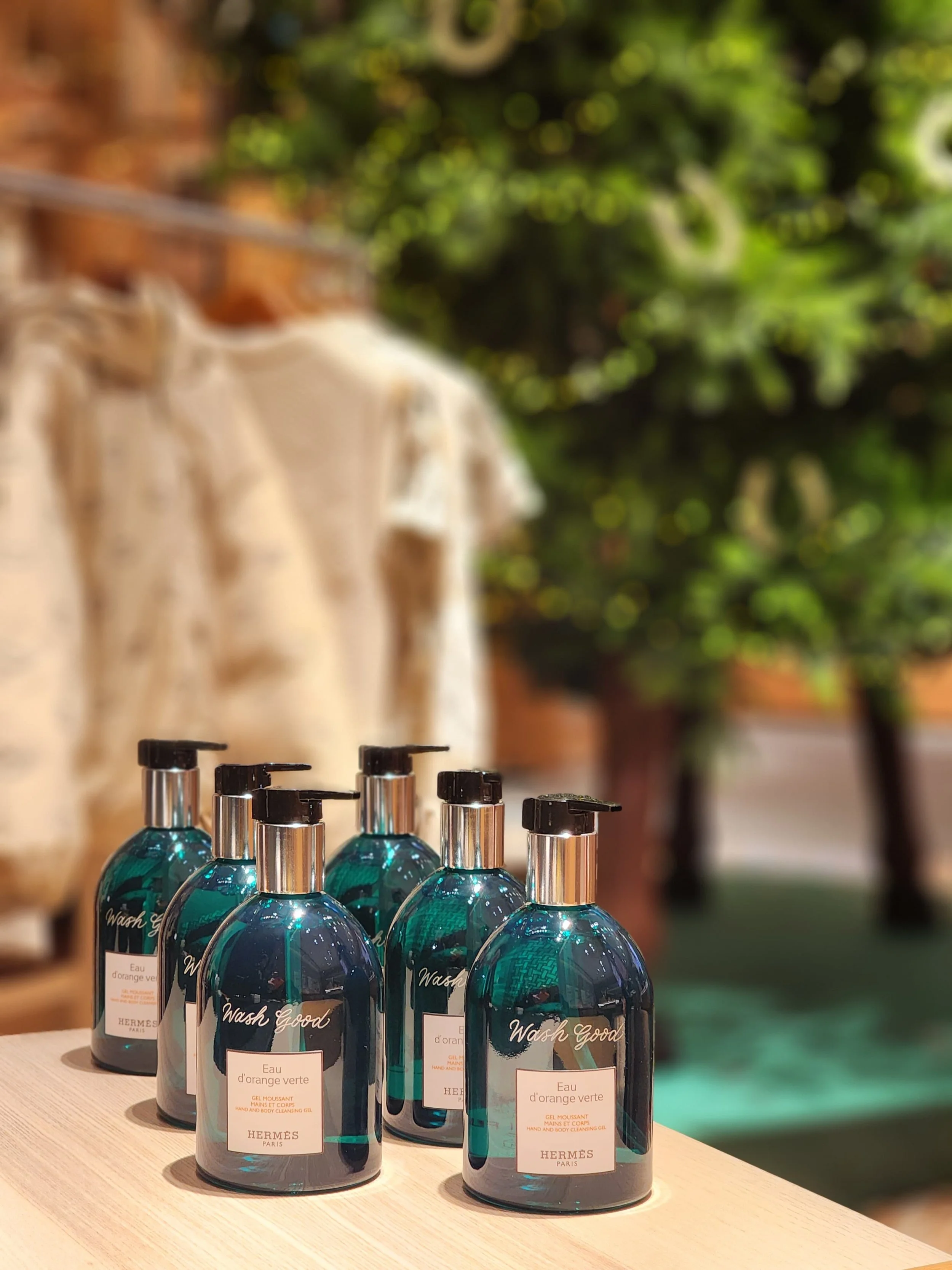 Six bottles of Hermes hand and body cleansing gel with pump dispensers on a wooden surface outdoors, blurred green trees and a brick wall in the background.