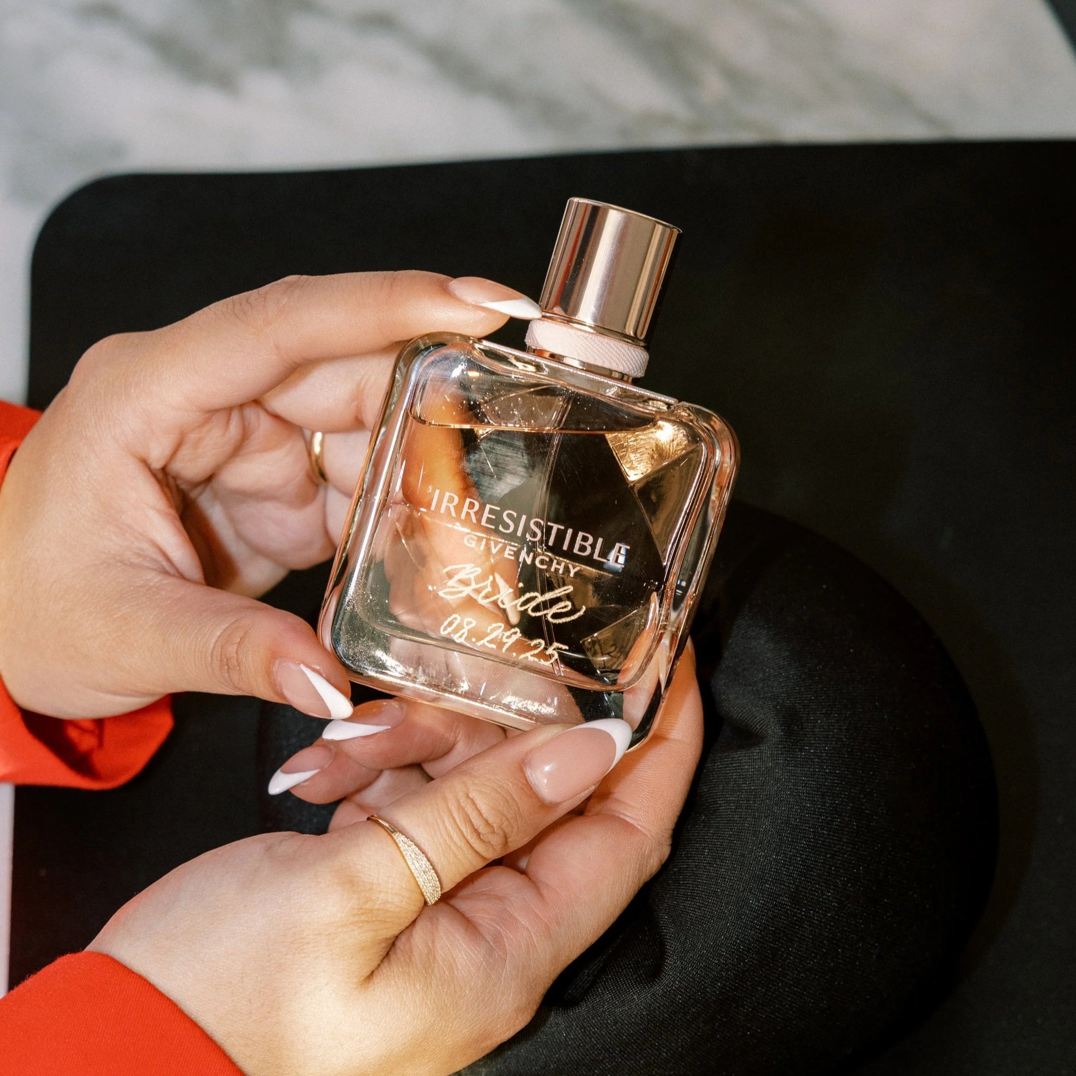 A hand holding a clear perfume bottle with a gold cap. The label reads 'Irresistible Givenchy' with a signature and date '08.29.25'. The person has manicured nails with a French tip design and is wearing a gold ring. The background is black and gray.