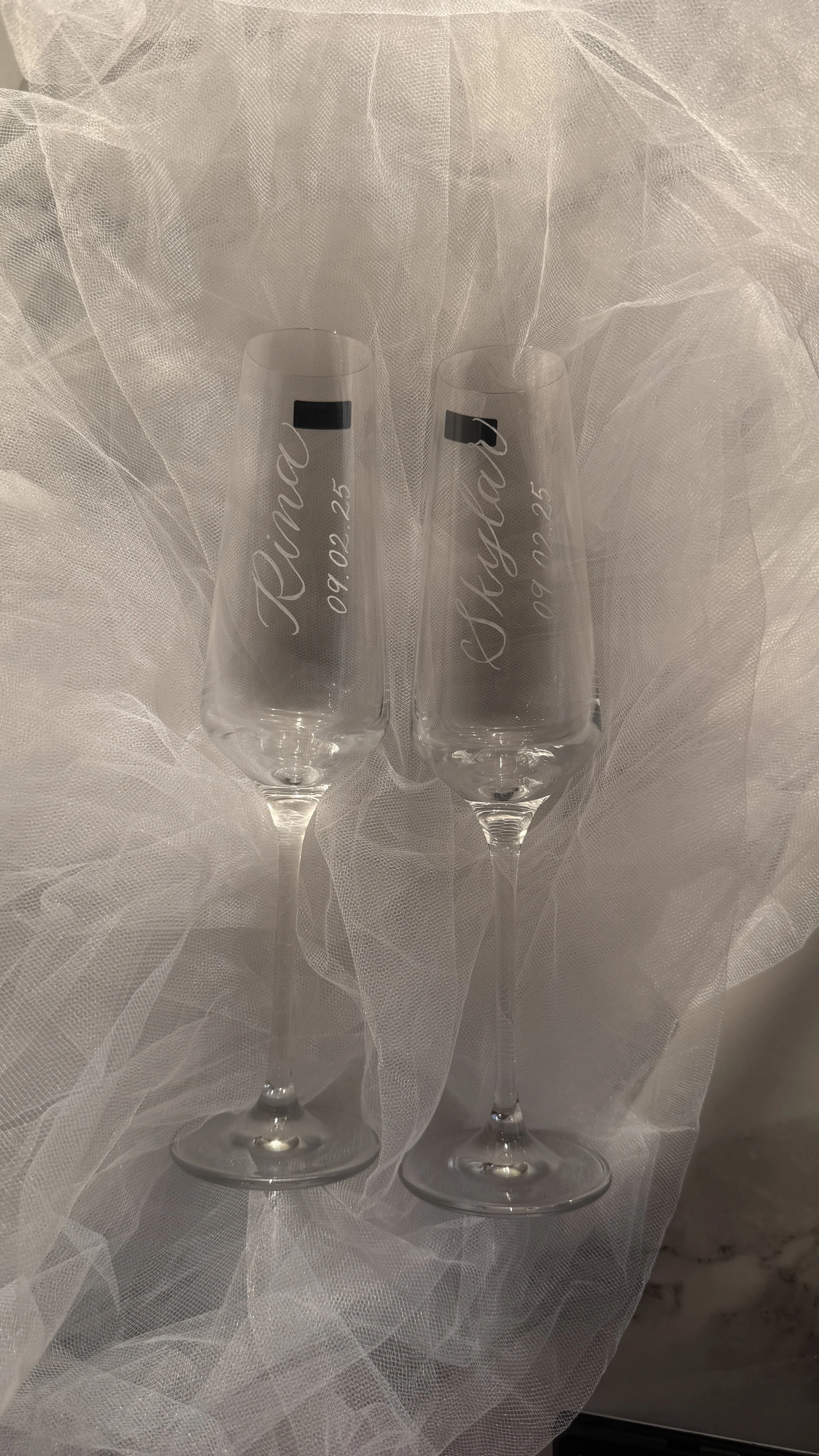 Two engraved champagne flutes with names and date, placed on white tulle fabric, likely for a wedding celebration.