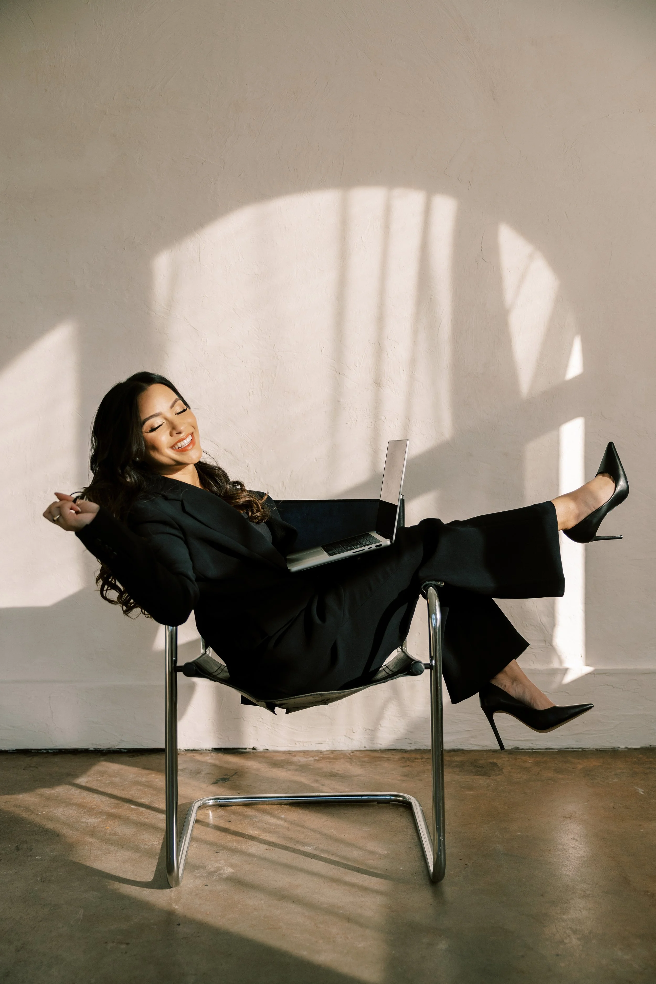 Woman in black business suit with high heels relaxing on a chair with a laptop on her lap, smiling with eyes closed, against a plain wall with sunlight and shadow.
