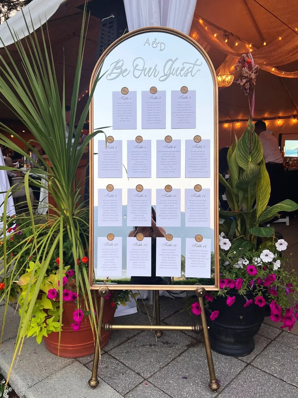 Wedding guest seating chart with a floral arrangement and large mirror at the top that says 'Be Our Guest' with tables numbered 1 through 15 listed below.