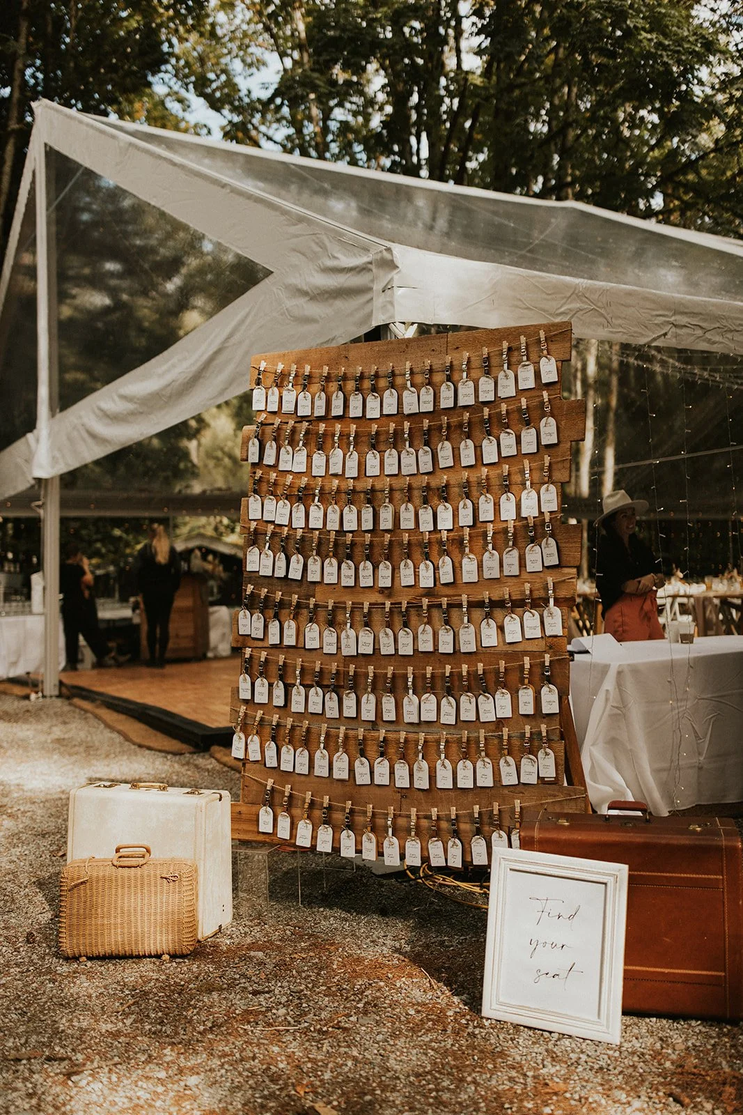 A wooden display board with multiple rows of white tags attached by clothespins at an outdoor event, with suitcases on the ground and a sign that reads 'Find your seat.' A white canopy tent and trees are visible in the background.