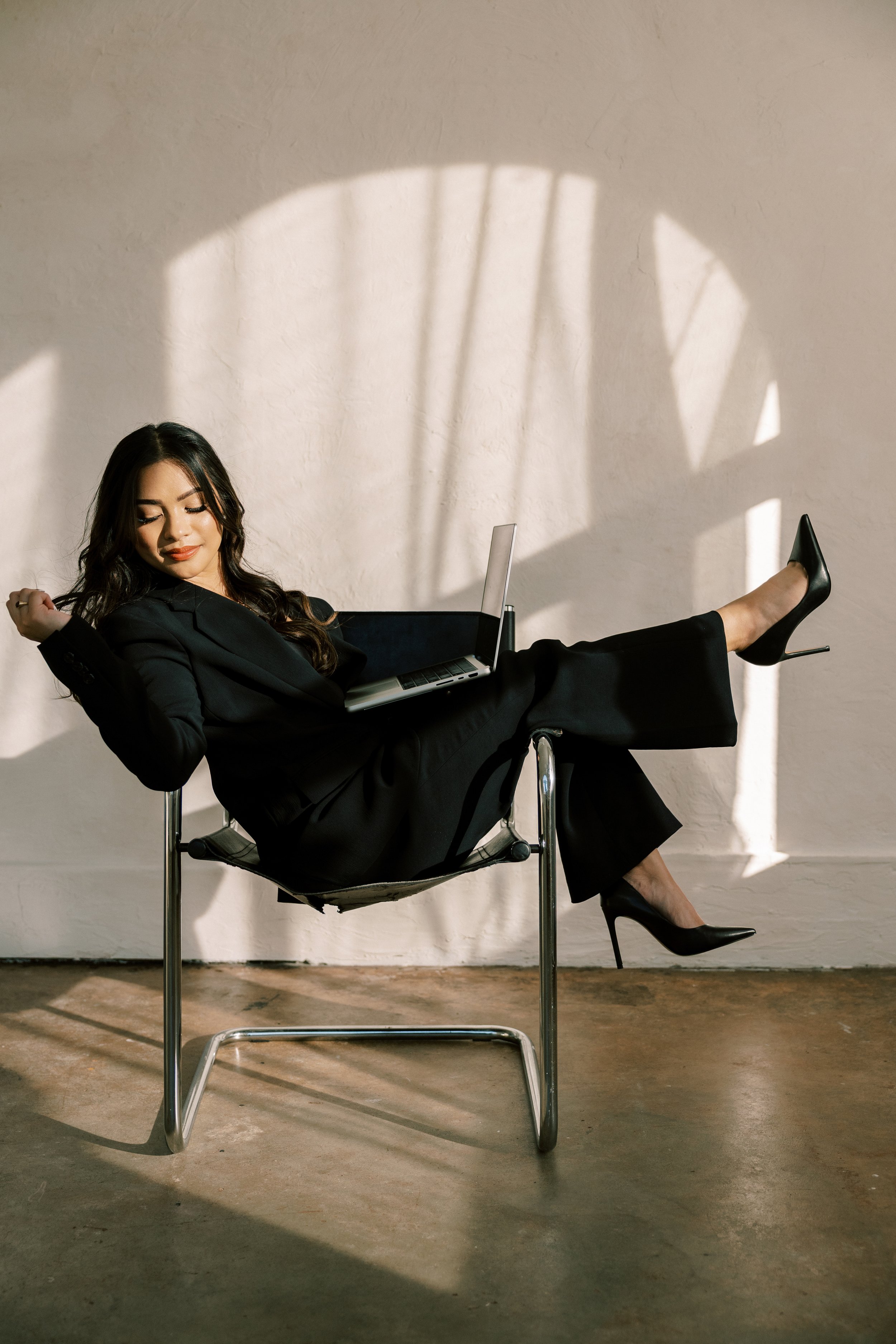 A woman in a black suit and high heels relaxes on a modern chair with her legs crossed, using a laptop, with sunlight and shadow on the wall behind her.