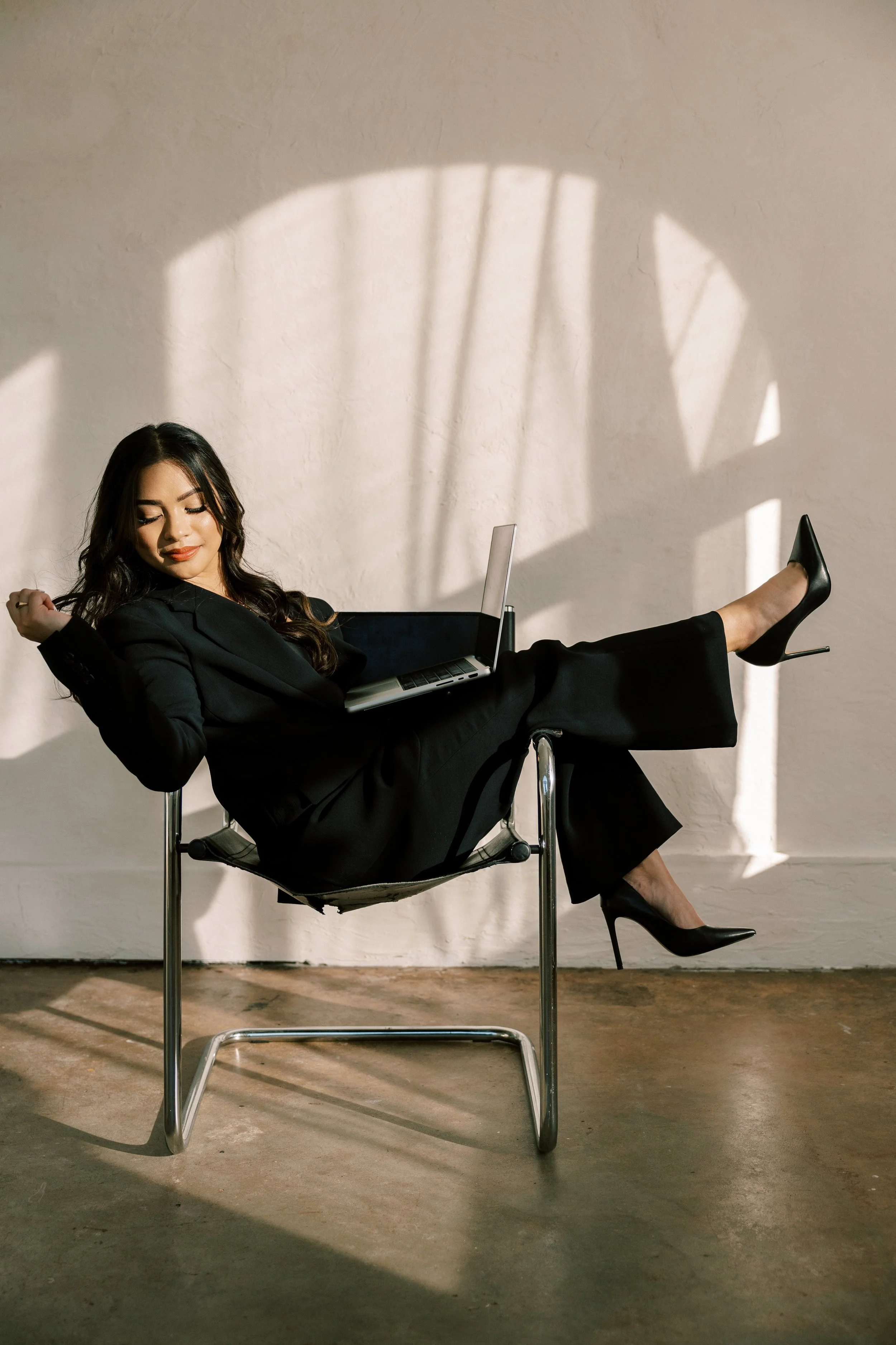 A woman in a black business suit and high heels relaxing with her legs up on a chair, holding a laptop on her lap, in a room with sunlight and shadows on the wall.
