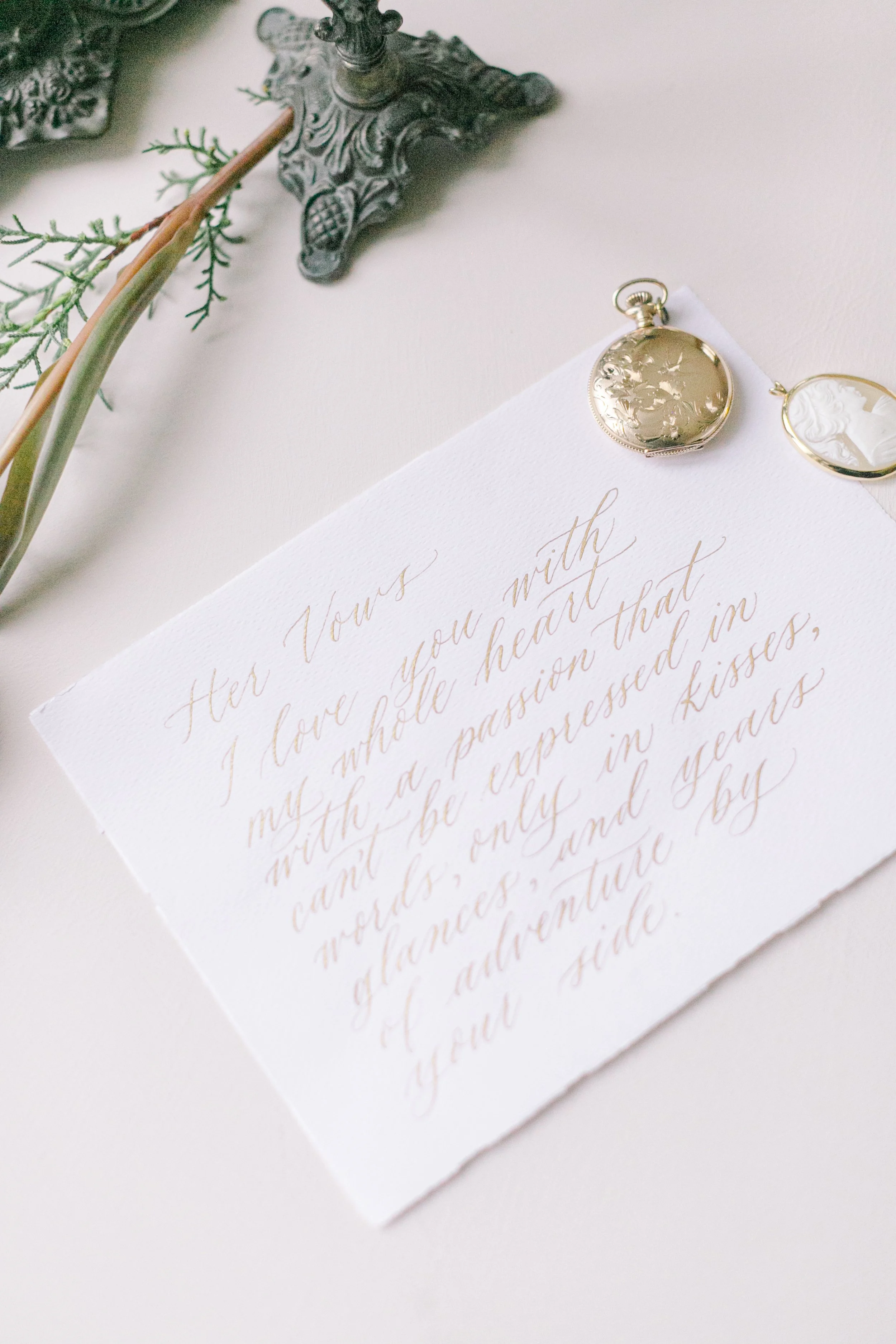 An elegant handwritten letter on textured white paper with gold ink, accompanied by a vintage pocket watch and cameo jewelry, and decorative objects including a metal stand and green plant stem.