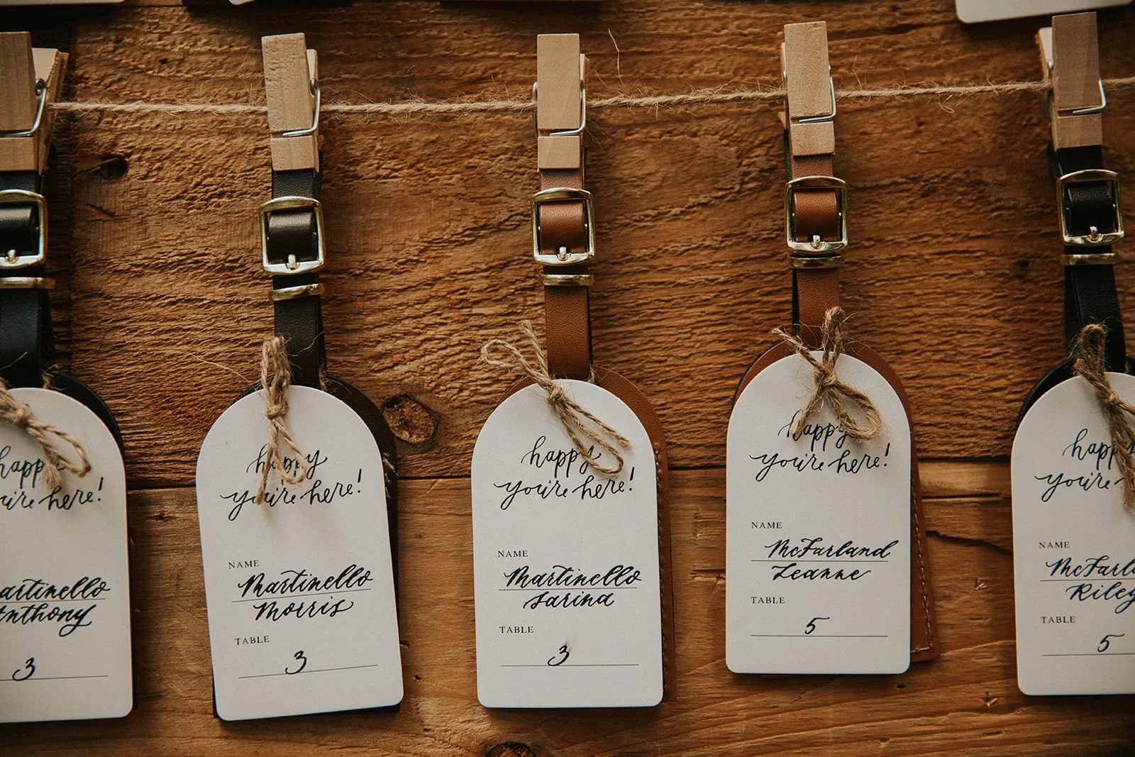 Wedding or event place cards hanging on a string with clothespins, on a wooden backdrop. The cards are labeled with guest names, table numbers, and feature a decorative handwritten font.