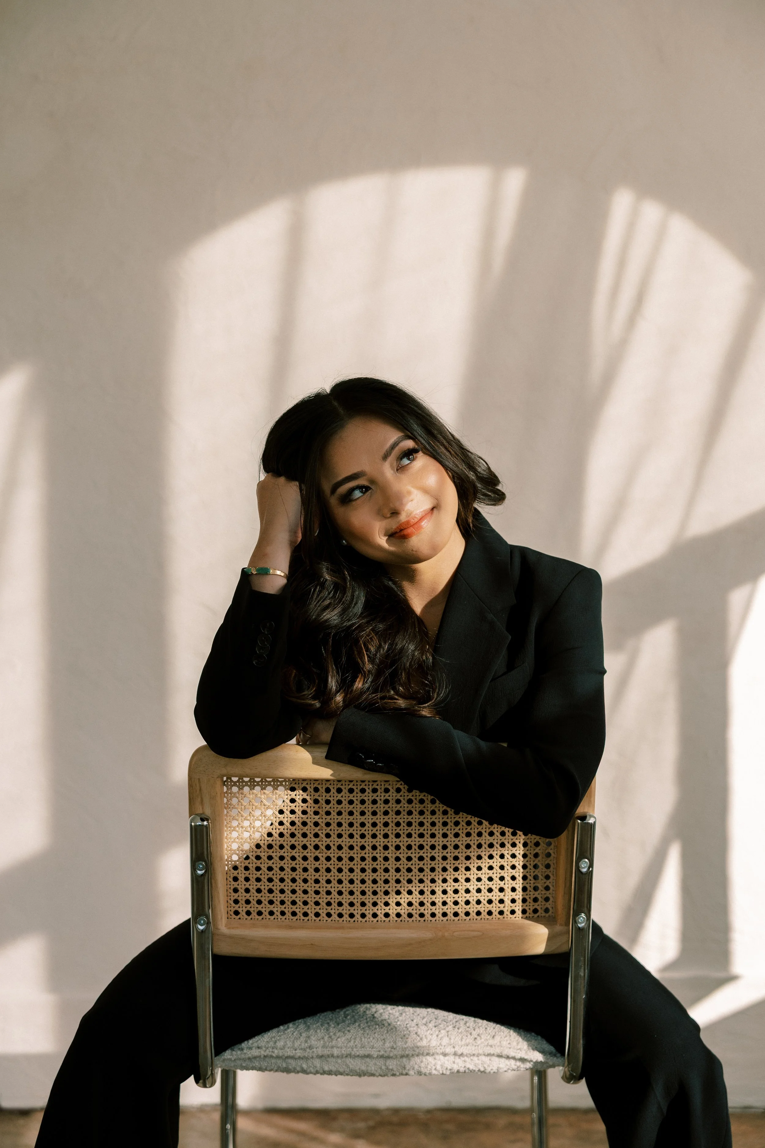A young woman with long dark hair, dressed in a black suit, sitting on a chair. She is resting her head on her left hand and smiling slightly, looking upward. Sunlight casts shadows on the wall behind her.