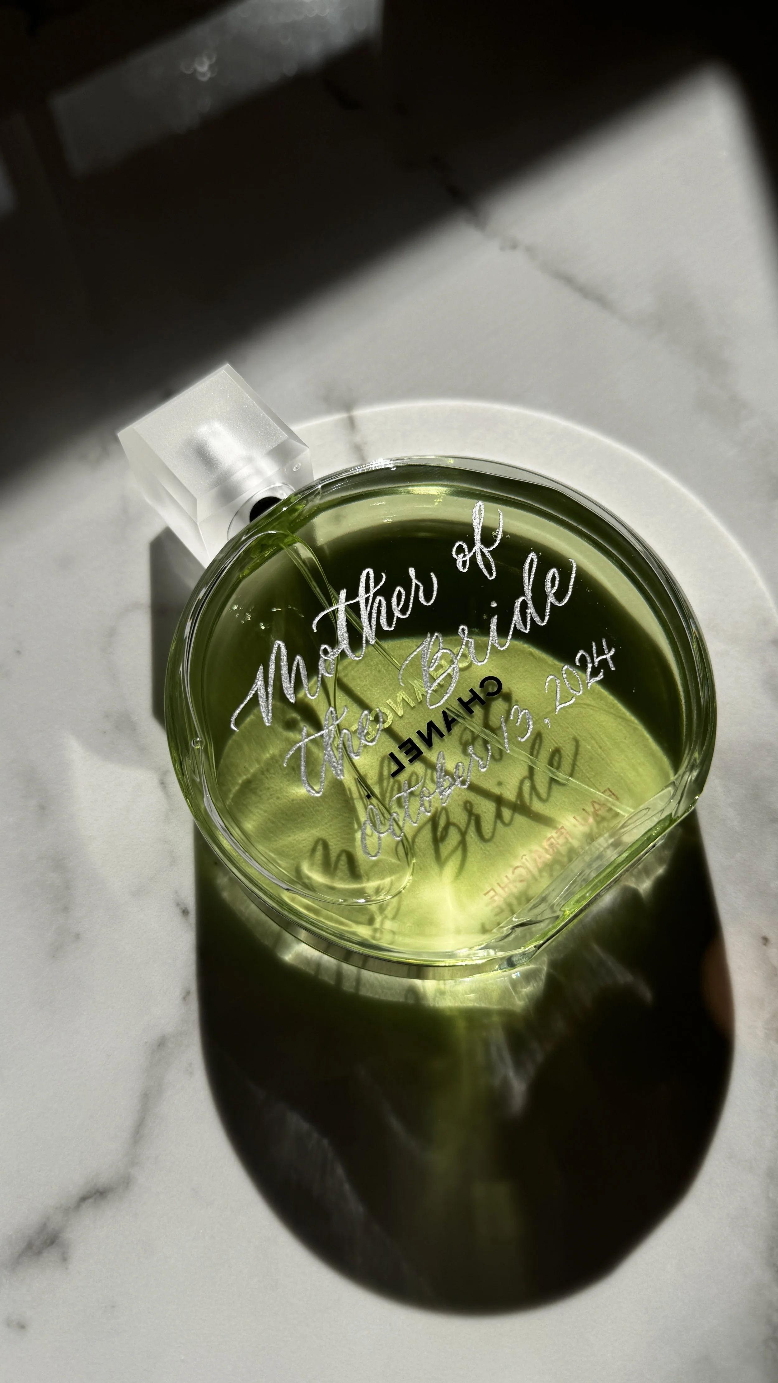 A glass container with yellow liquid, possibly a candle, sitting on a white surface. The glass has engraved text that reads, 'Mother of the Bride, October 13, 2014, Chelsea and Brad.'