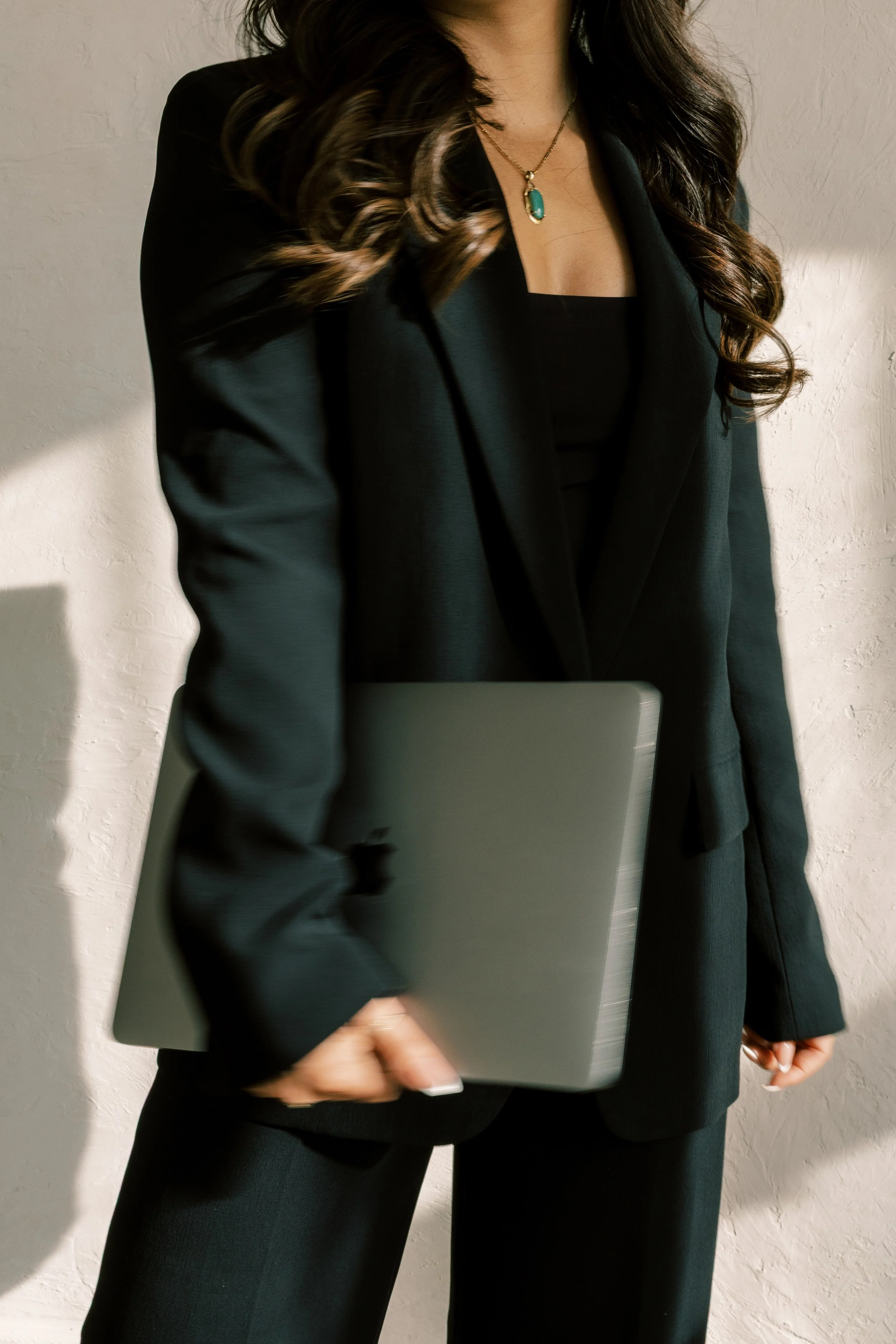 Woman in black suit holding closed laptop, standing against white textured wall.