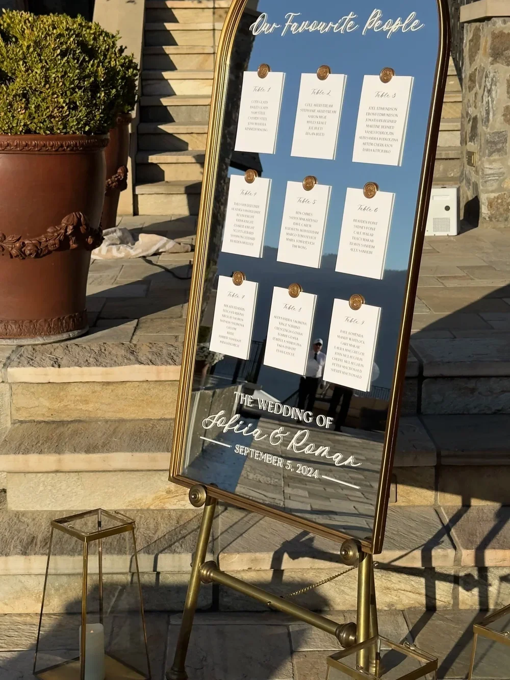 A gold-framed wedding seating chart on a glass board with handwritten script, displaying table assignments for guests at the wedding of Sofia and Roman on September 5, 2024. The board is outdoors near steps, with a large potted plant and lanterns nearby.