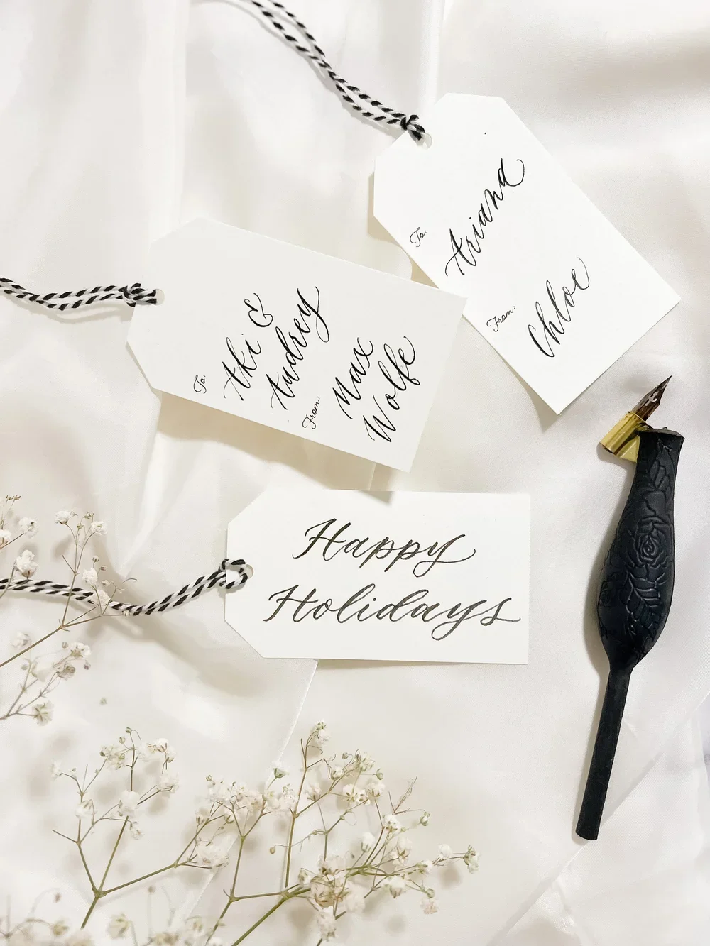 White holiday gift tags with black cursive writing, a black and gold ink pen, and small white flowers on a white surface.