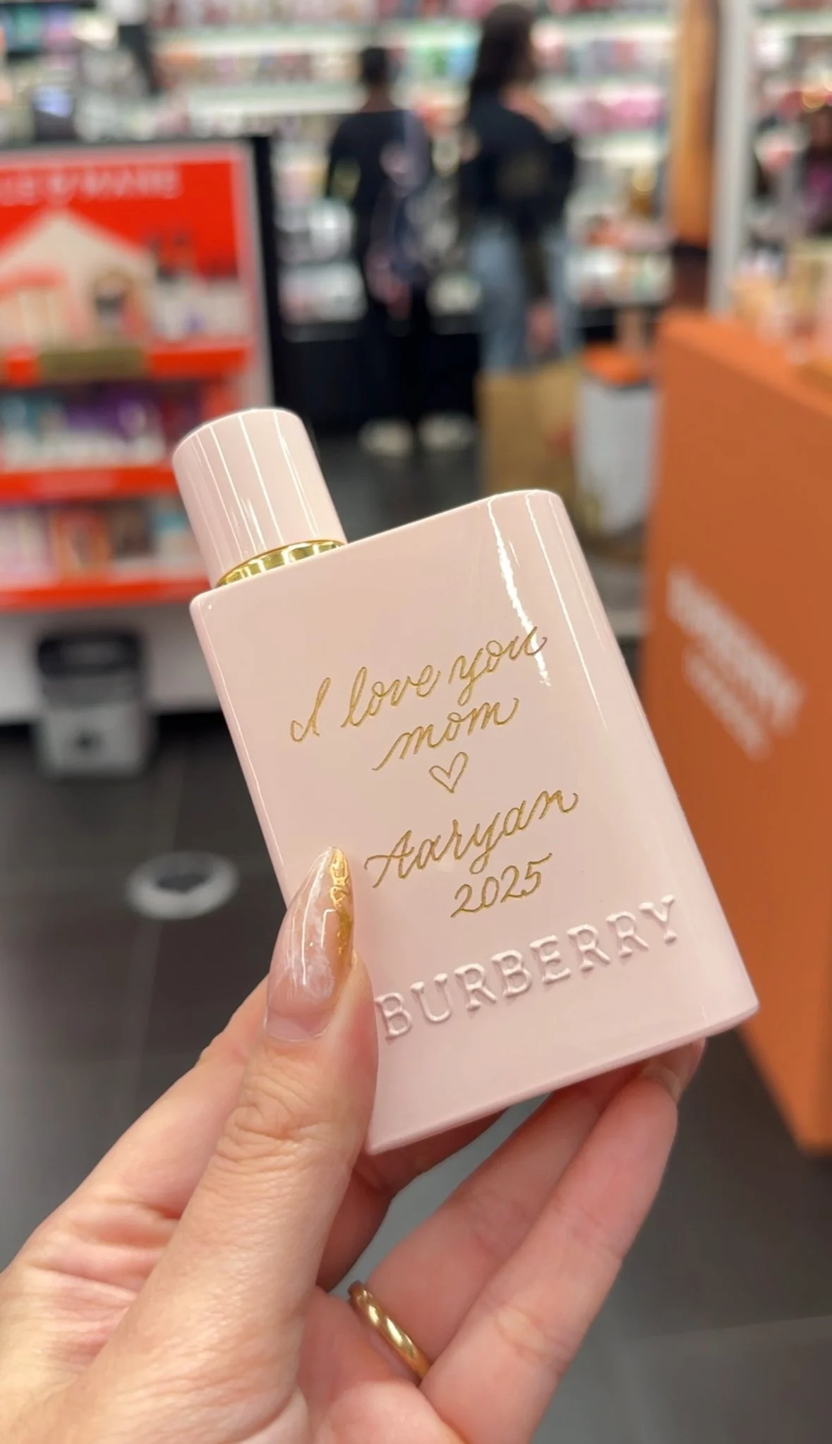 A pink Burberry perfume bottle with gold text reading 'I love you mom, Aaryan 2025' held in front of a blurred store background with two women shopping.