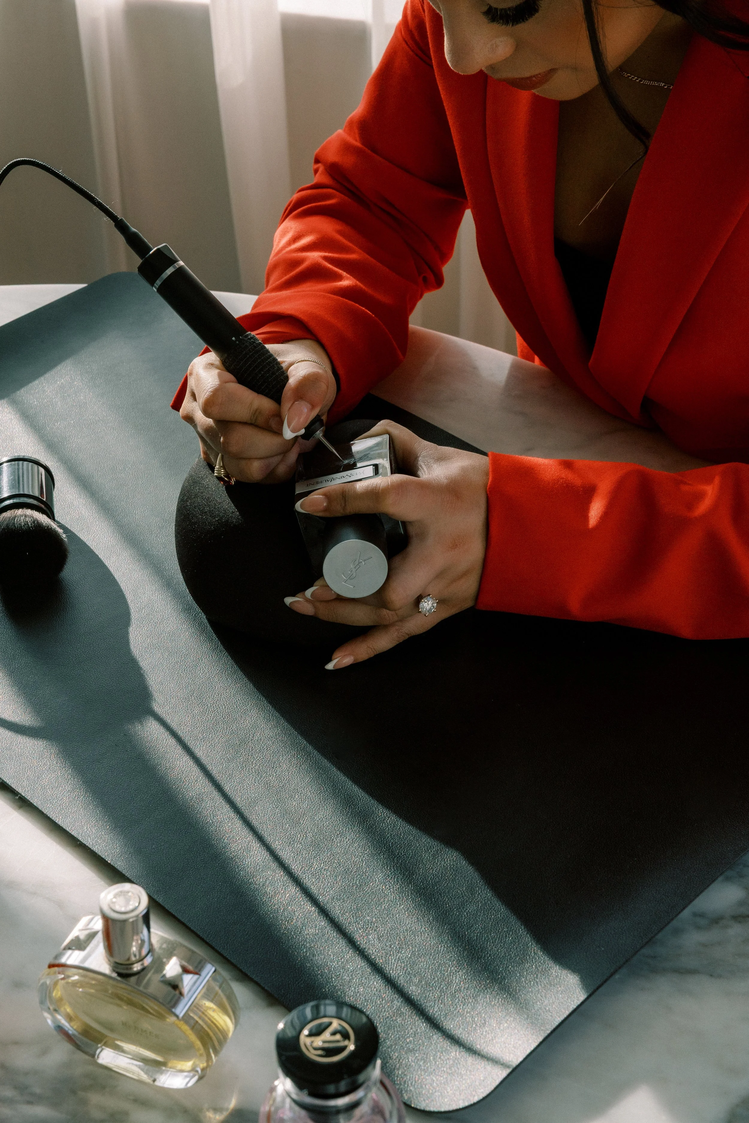 A woman in a red blazer is getting a tattoo of her thumbprint on her finger, using a tattoo machine.