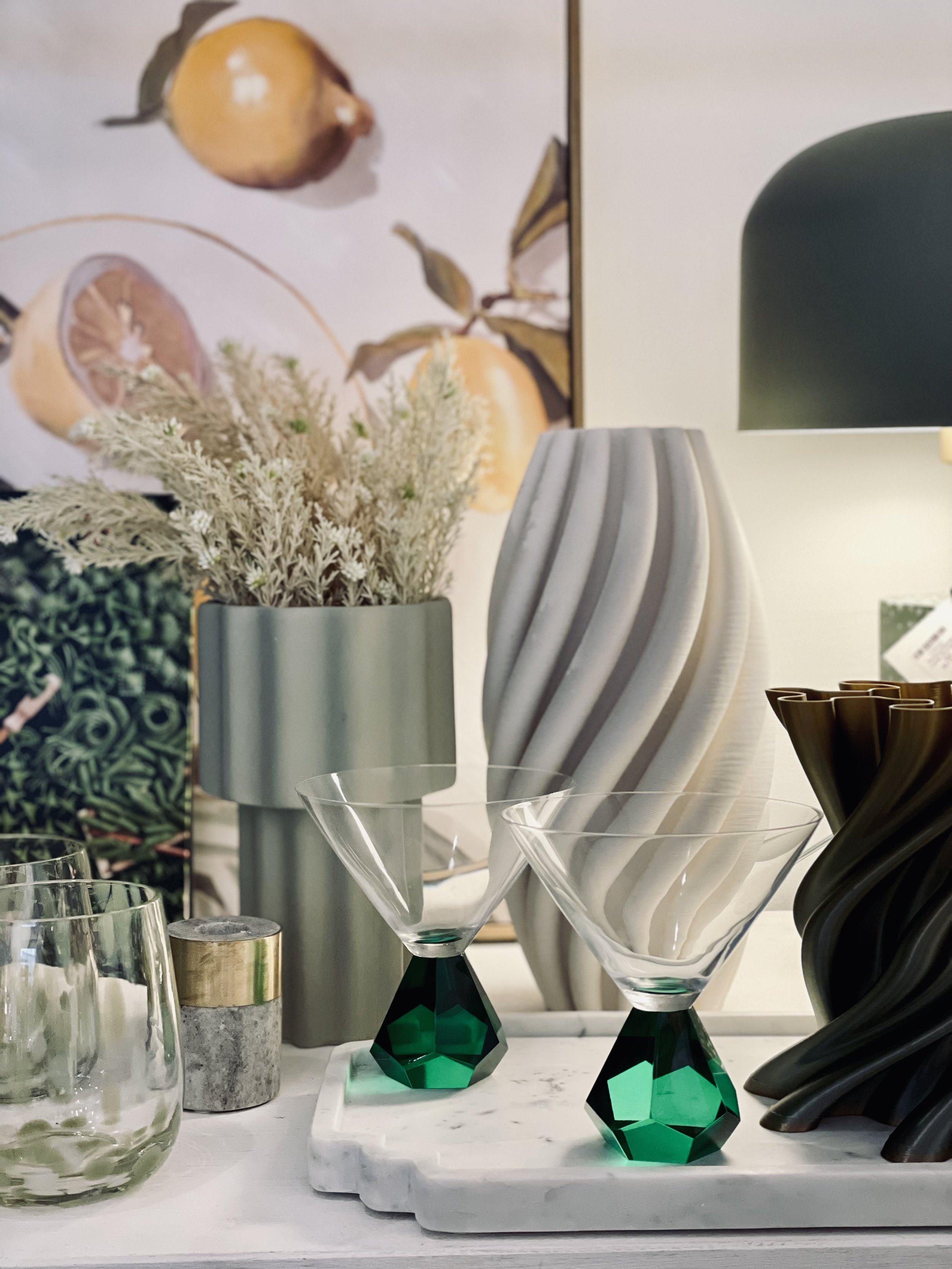 A decorative arrangement featuring art pieces and home decor items. A wall art piece with lemons is in the background, alongside a large gray vase with white dried flowers. Two martini glasses with green geometric bases are on a white tray. There's also a cylindrical candle and two other vases with spiral designs, one gray and one bronze. A green lamp is partially visible on the right.