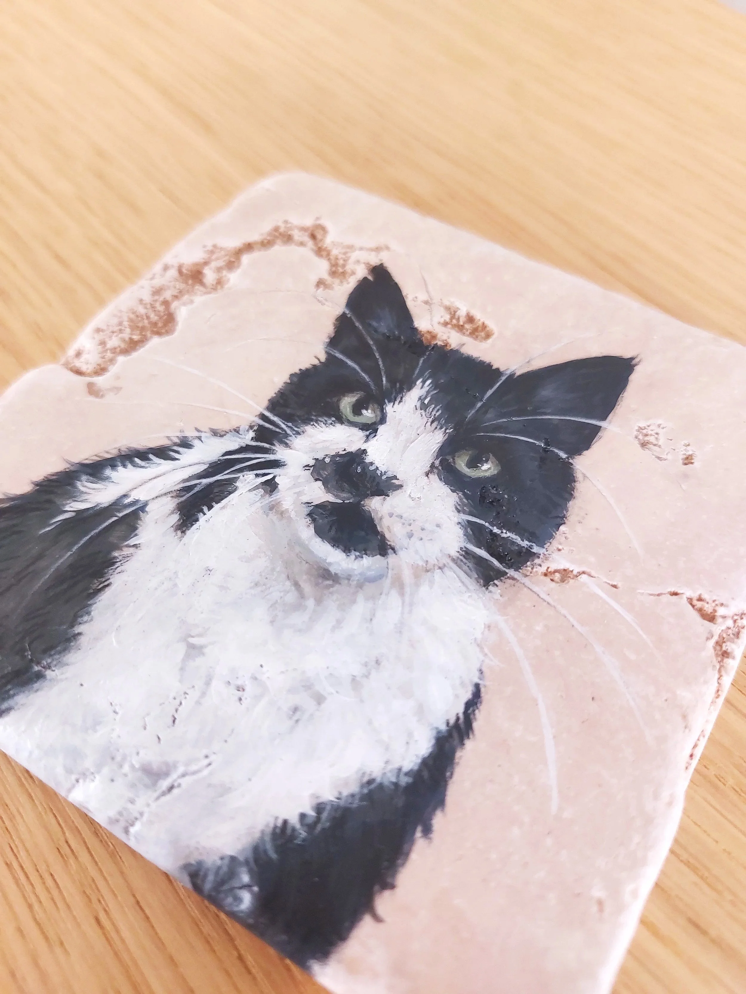 Photo of a cat painted on a ceramic tile or piece of pottery, with a black and white fur pattern and green eyes.