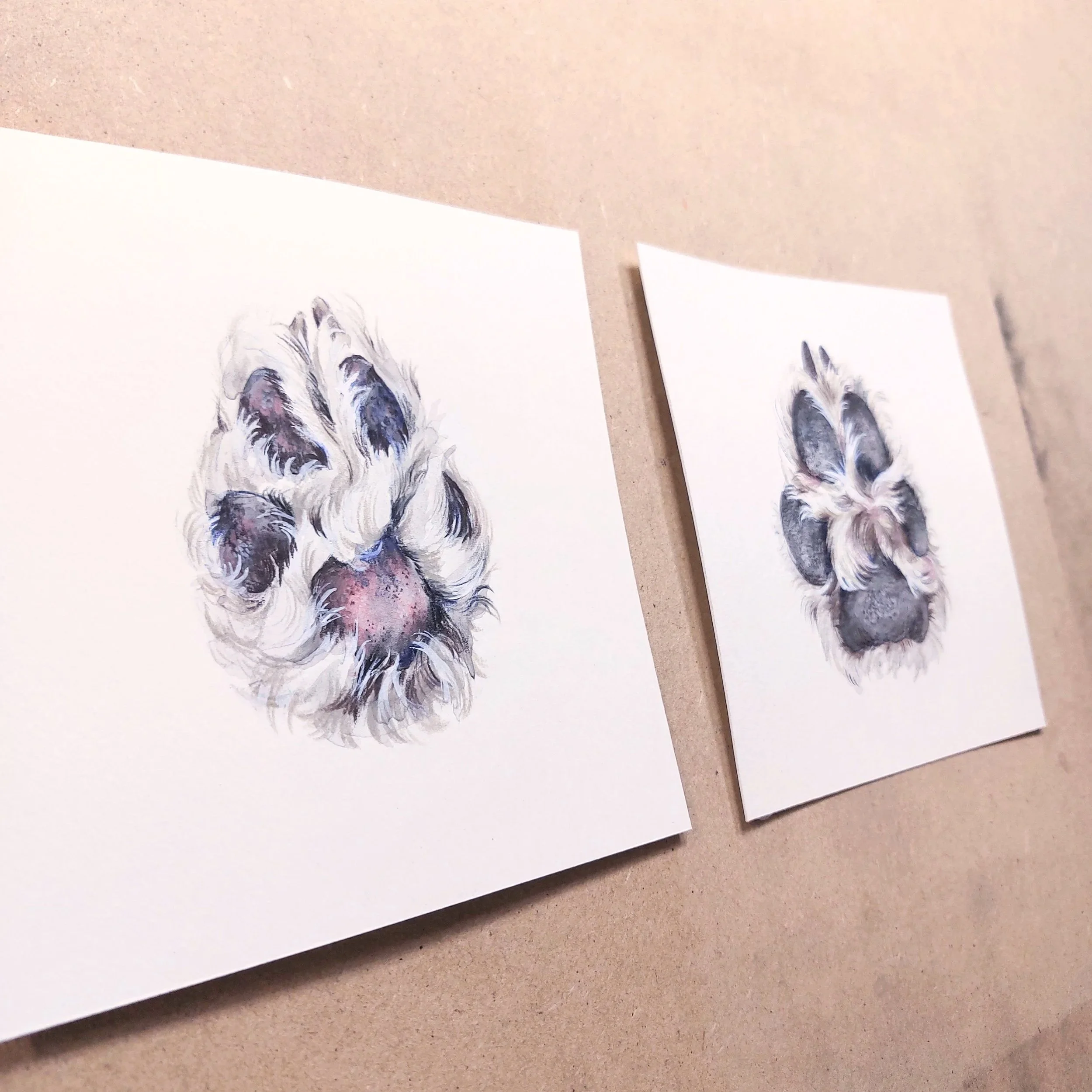 Two watercolor paintings of a dog’s paw prints on white paper, displayed on a beige surface.