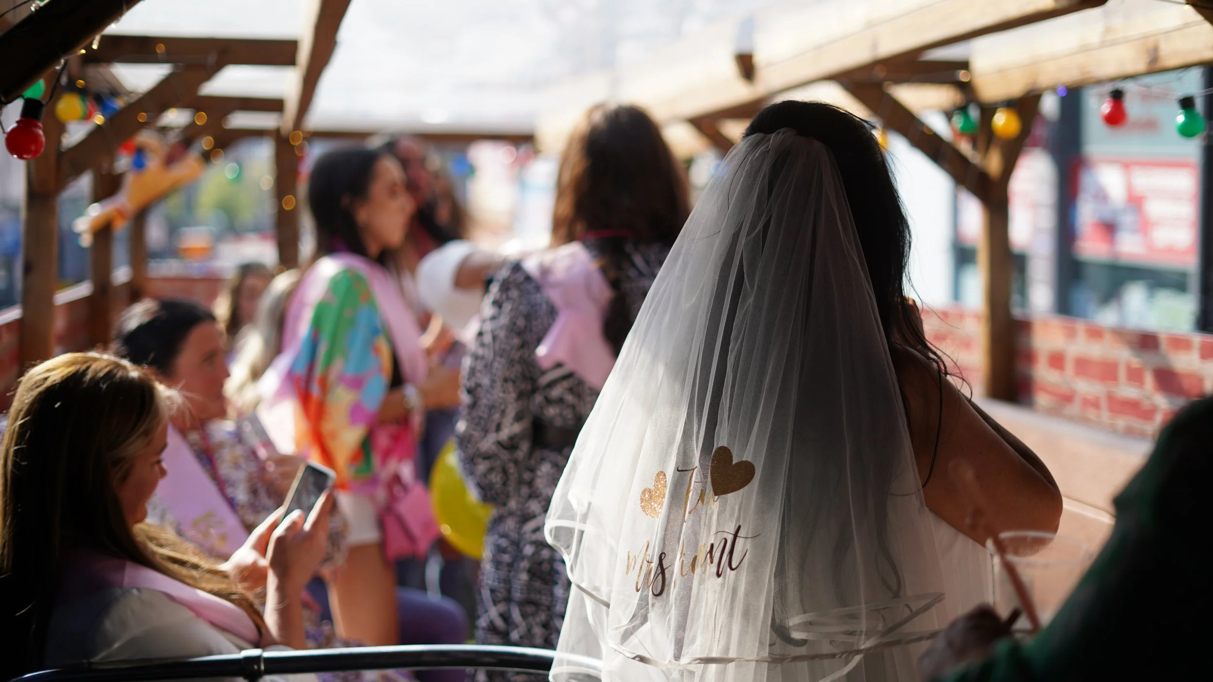 A bride with a veil stands outside a decorated venue, surrounded by women, some of whom are taking photos and talking.