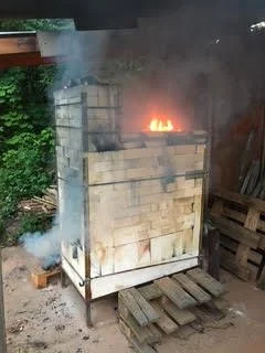 Holzbrandofen während des Brandes, Flammen schlagen aus der Ofendecke, Rauch zieht über den Garten