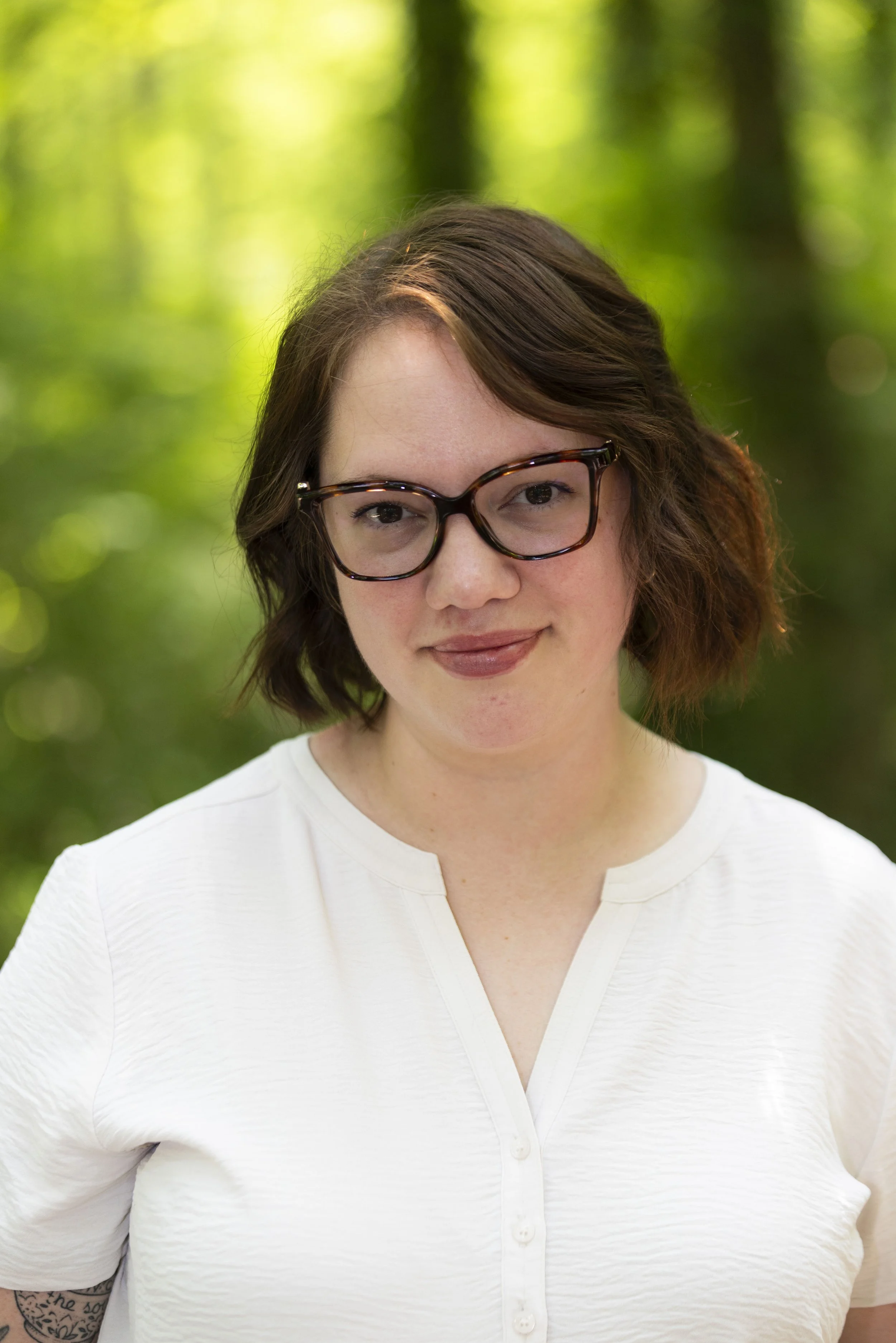 Portrait of a smiling woman with shoulder-length brown hair, wearing a white blouse, and glasses outside with greenery in the background.