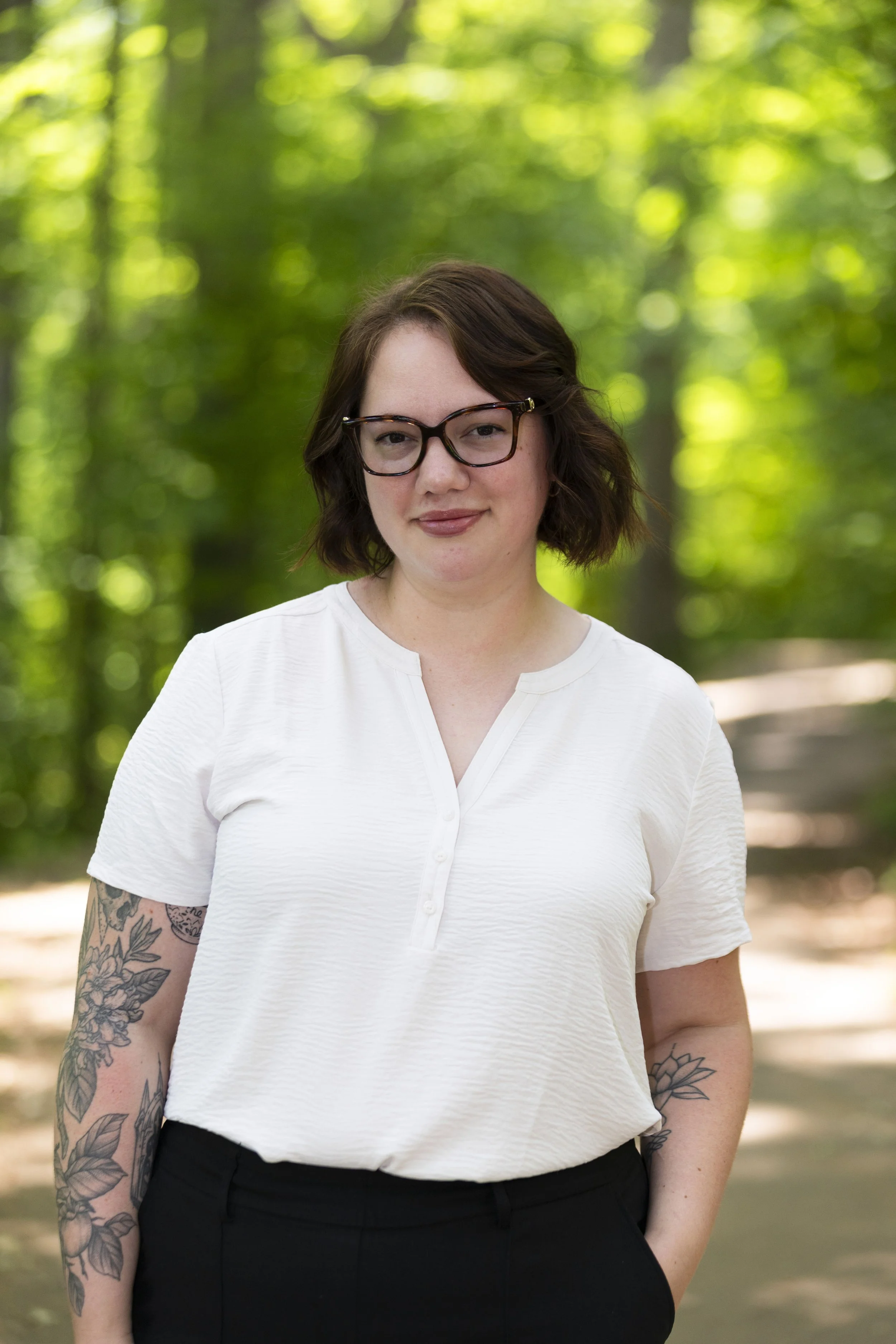 A woman with brown hair, glasses, and tattoos on her arms, standing outdoors in a wooded area.