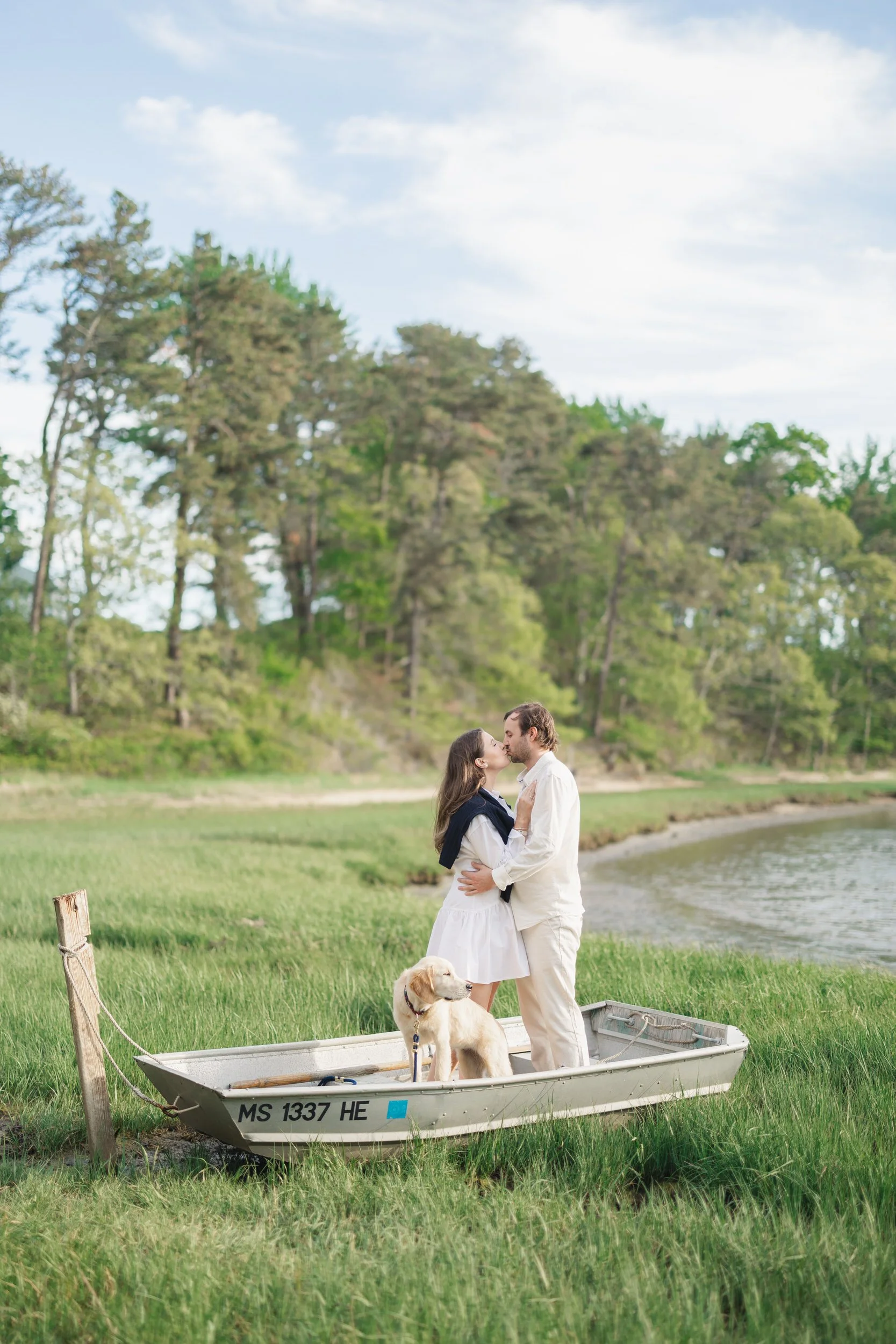 Couple standing on a boat surrounded by tall green grass during a Cape Cod engagement session in Chatham Cape Cod, with a golden retriever puppy and soft coastal light.