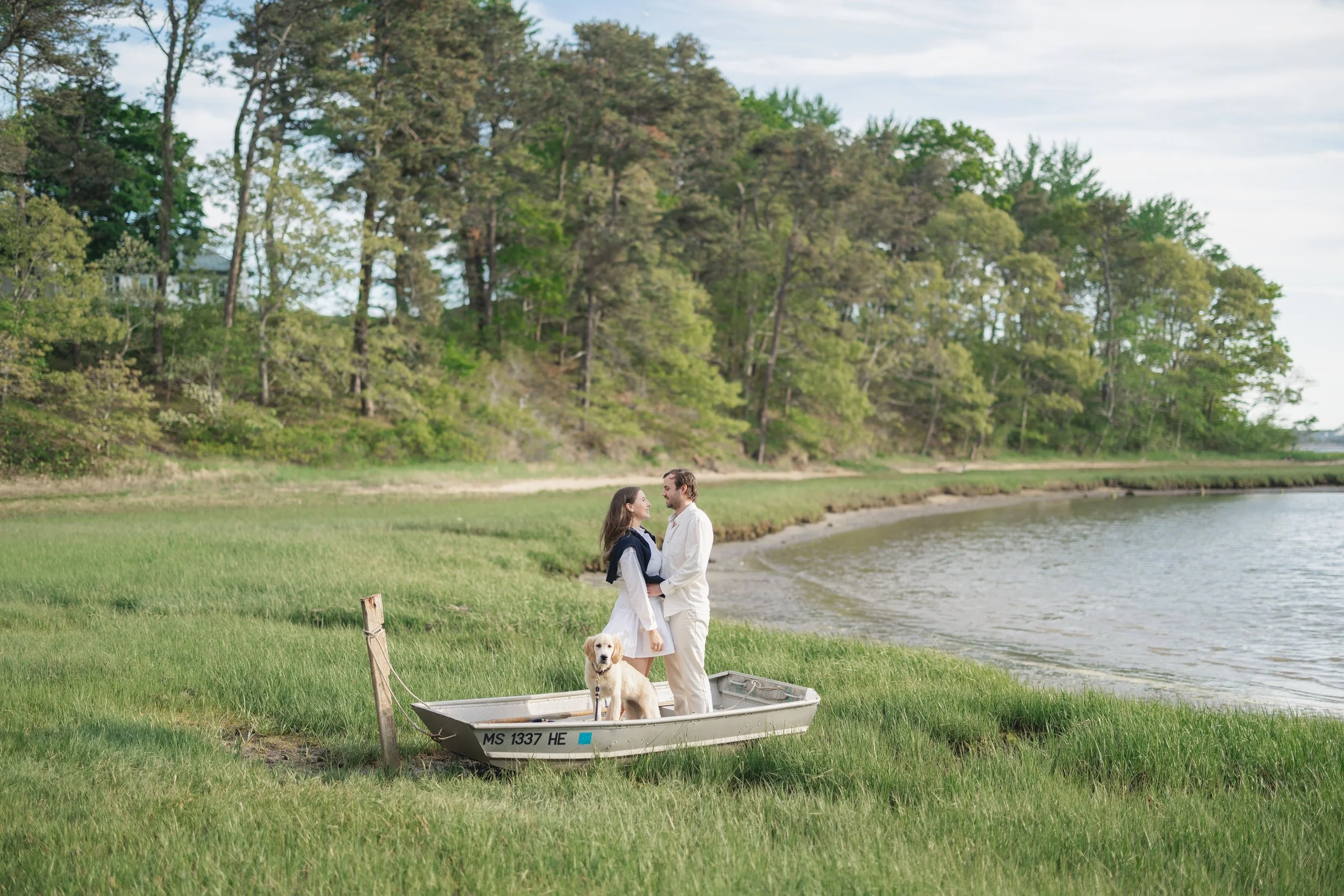 Couple standing on a boat surrounded by tall green grass during a Cape Cod engagement session in Chatham Cape Cod, with a golden retriever puppy and soft coastal light.