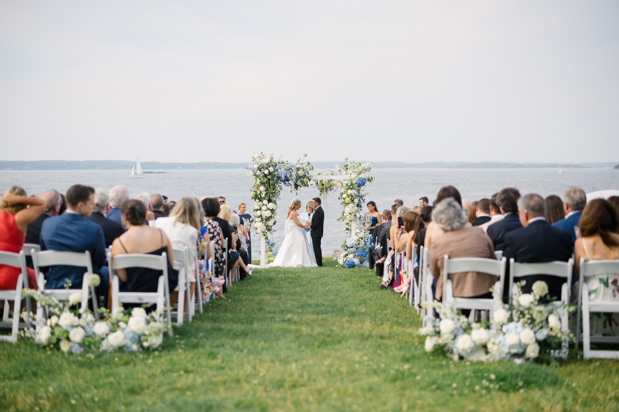 Bride and groom exchanging vows during a waterfront ceremony in Marblehead, Massachusetts with sailboats in the background.