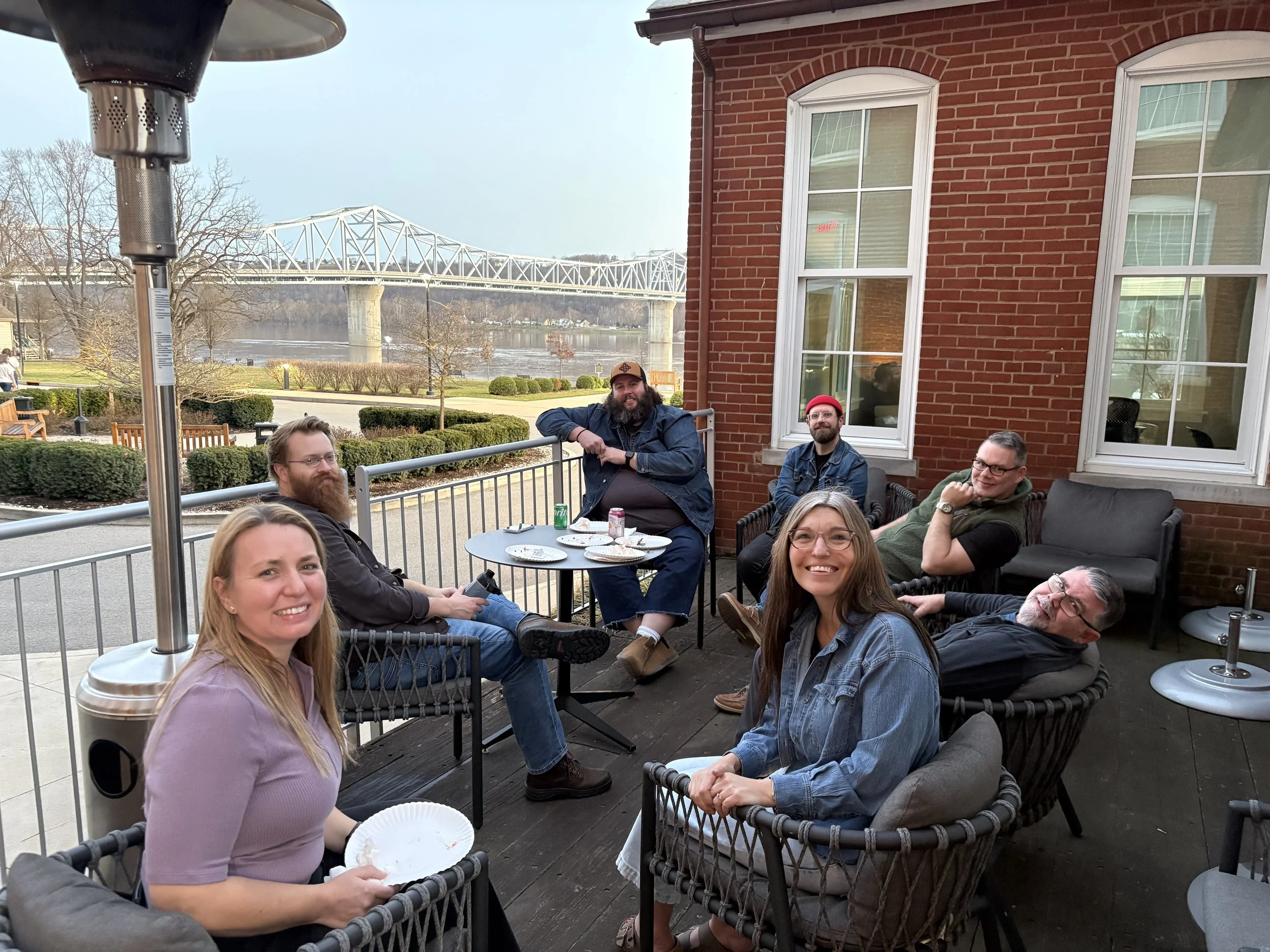 A group of people sitting on a patio back deck enjoying food and drinks. In the background, a river, bridge, and trees are visible.