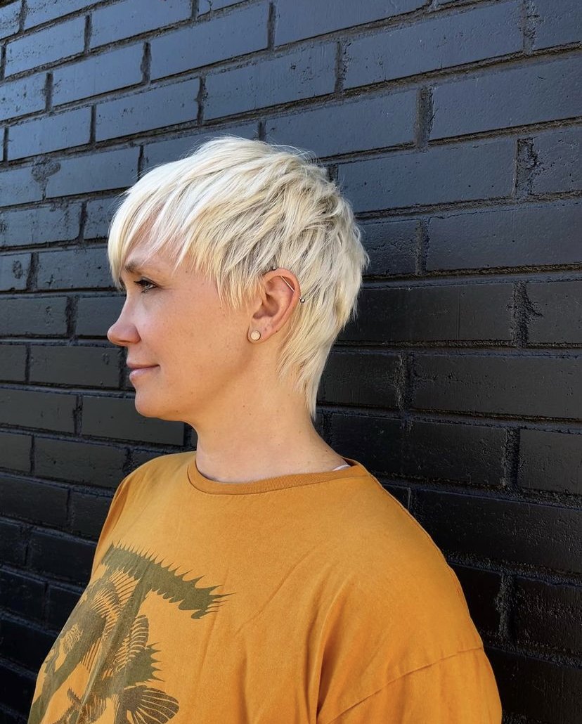 Person with short blonde hair wearing an orange shirt standing against a black brick wall.