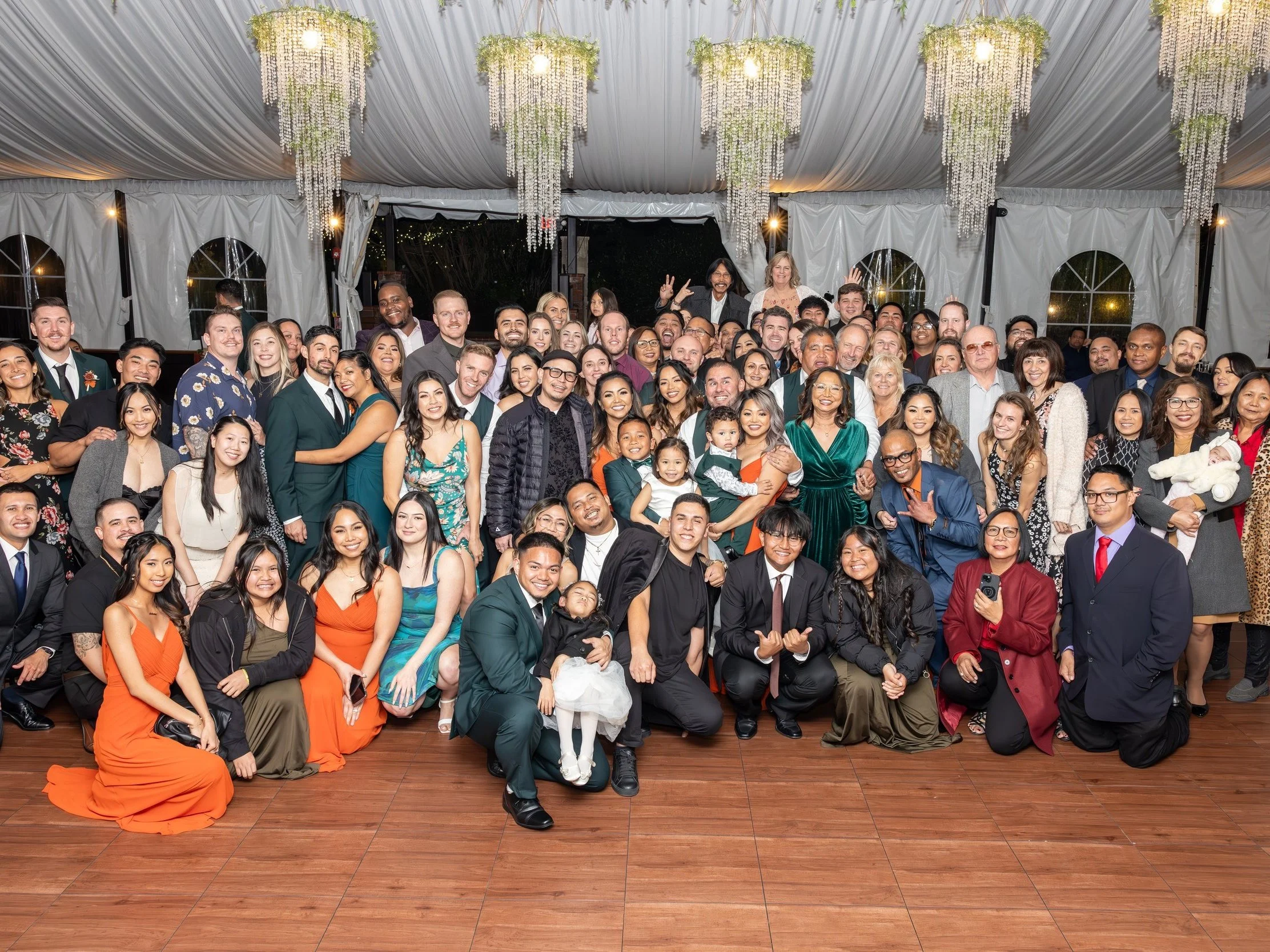 Large group of diverse people posing for a group photo at an indoor event under decorated ceiling with hanging floral arrangements, some people sitting, others standing, smiling and celebrating.