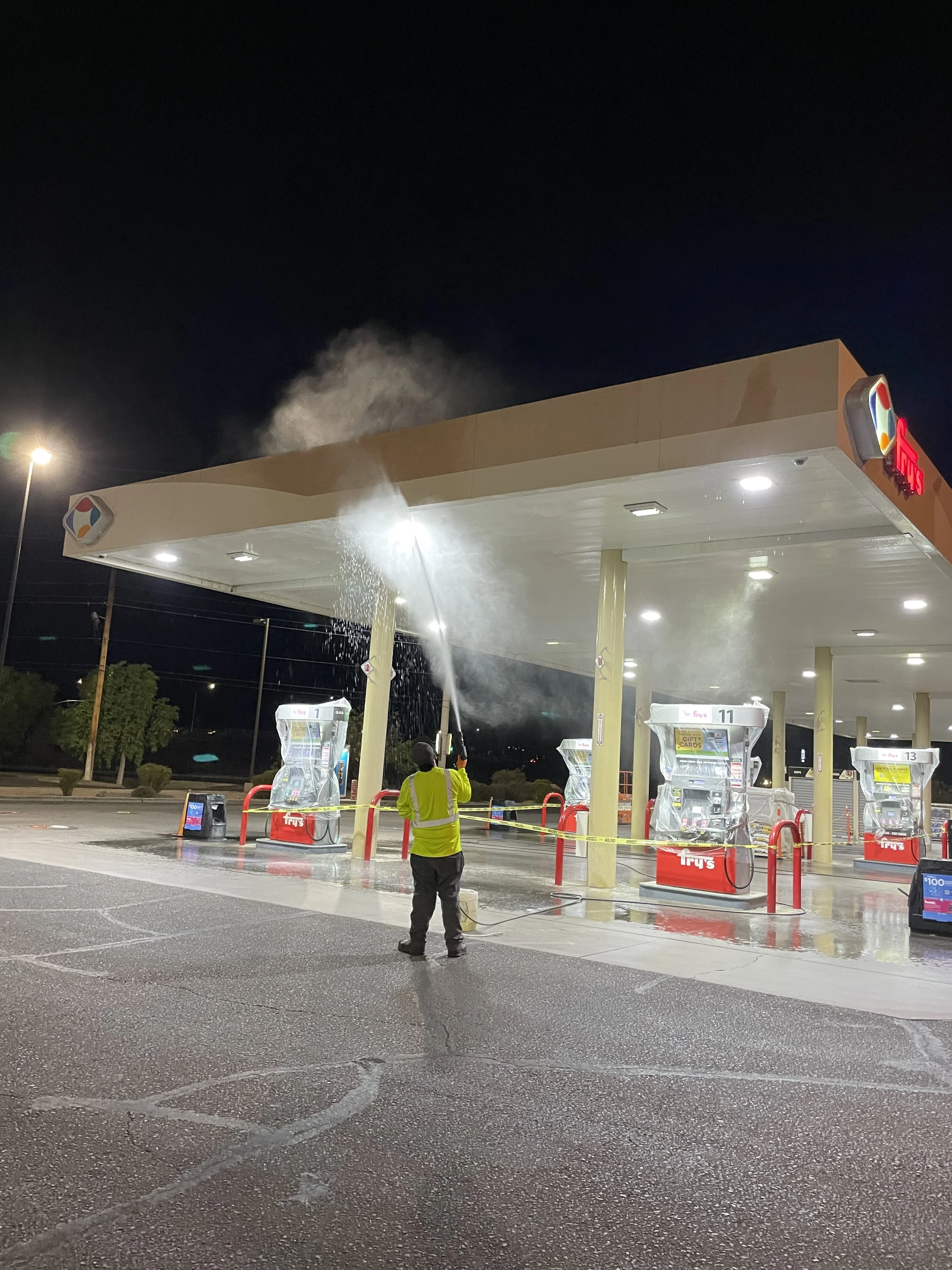 A person cleaning a gas station pump at night with a high-pressure water hose, with steam rising from the equipment, under bright overhead lights.