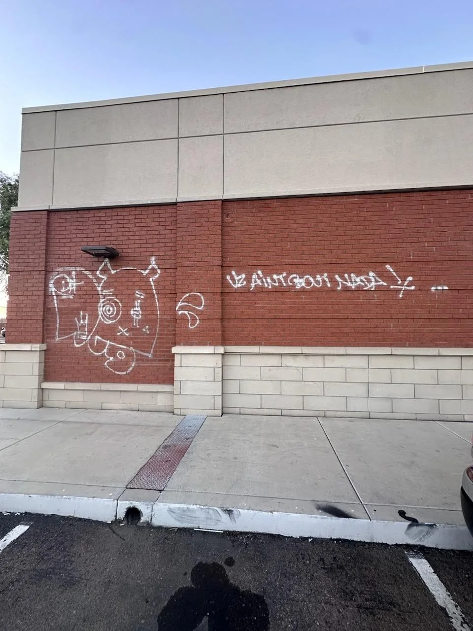 Street view showing graffiti on a red brick wall of a building, with a cartoonish bull's head and the phrase "BE ANGRY BUT NOT !!!" written in white spray paint.