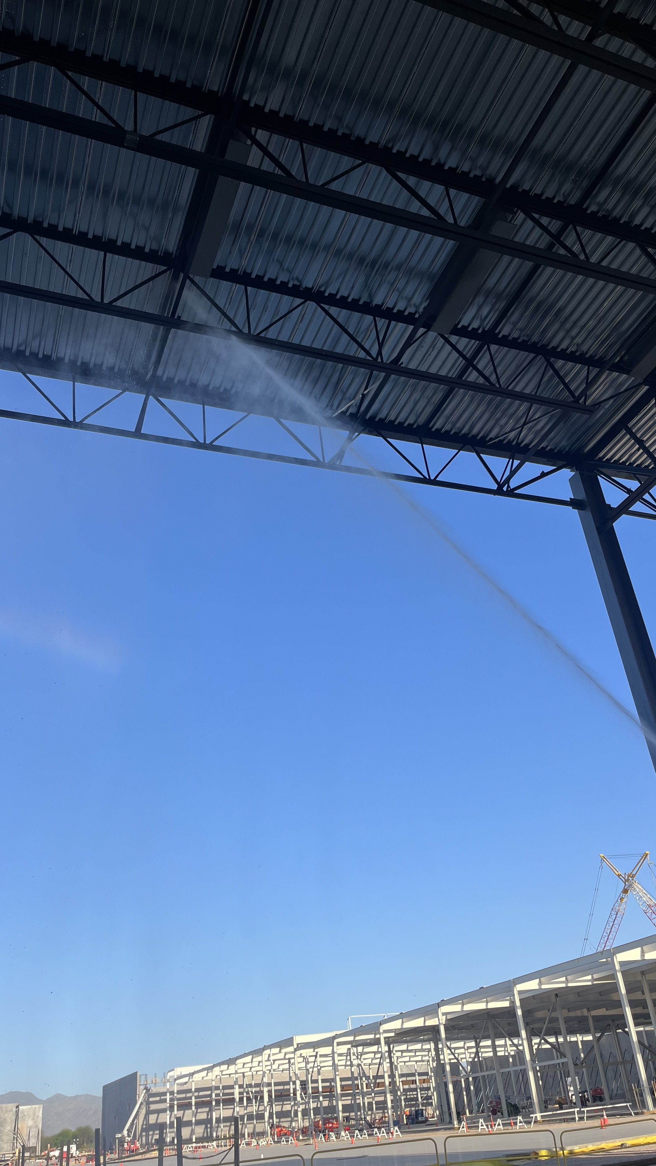 Under construction stadium with a large steel framework, bright blue sky, and construction cones and equipment.