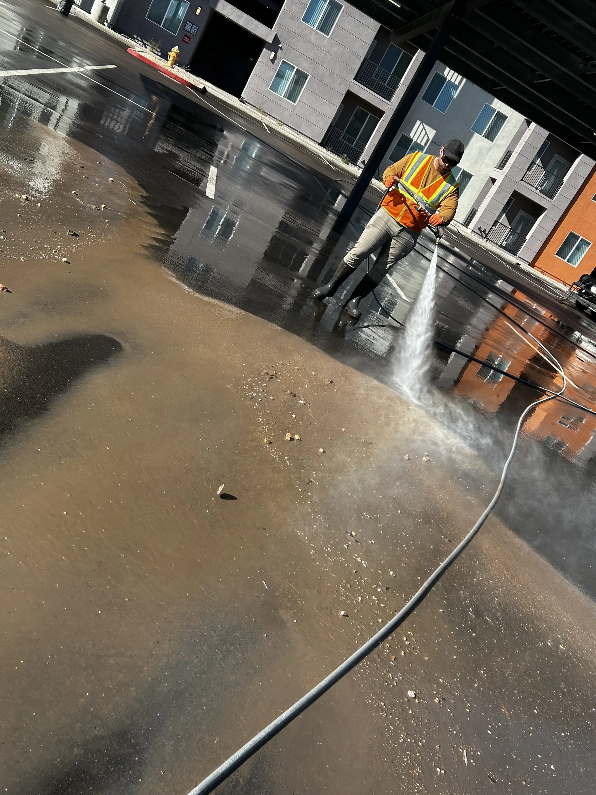 A worker in a yellow and orange safety vest and boots is using a power washer to clean a parking lot, with residential buildings in the background.