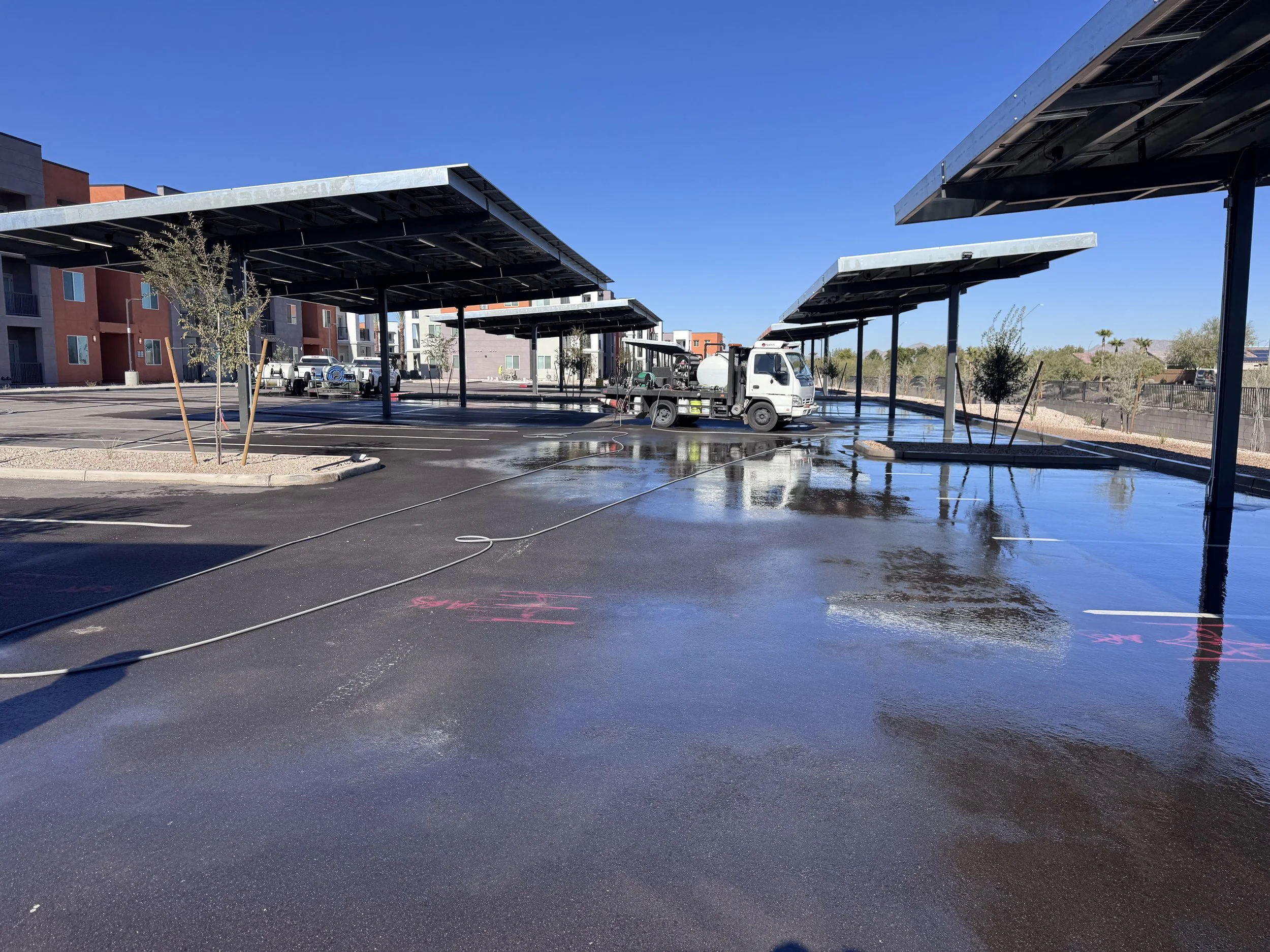 Parking lot with solar panel covered parking structures and a water truck spraying water onto the pavement on a sunny day.