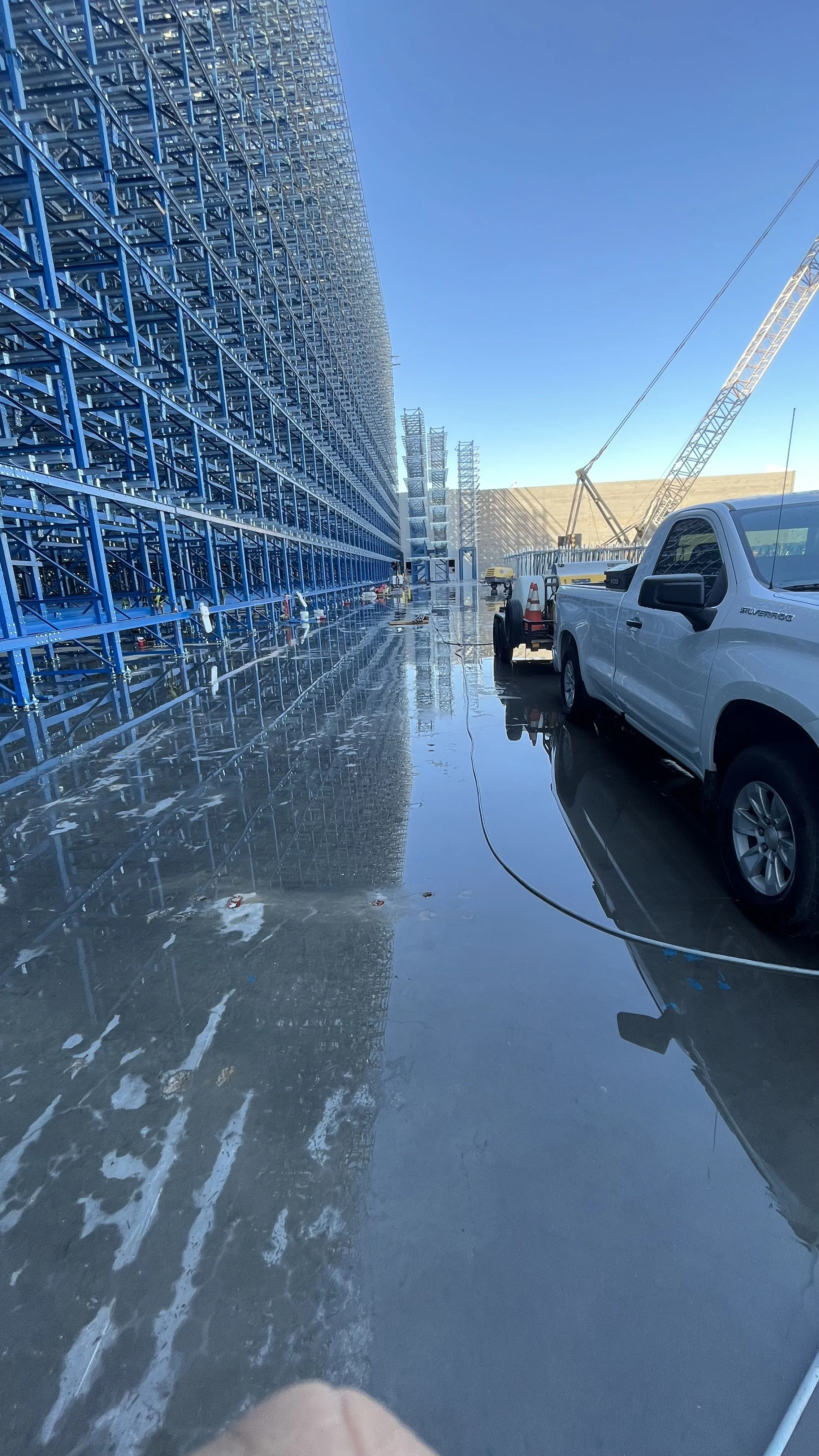 Construction site with a tall blue steel framework building on the left, a white pickup truck and construction equipment on the right, and a blue sky above. The ground is wet and reflective.