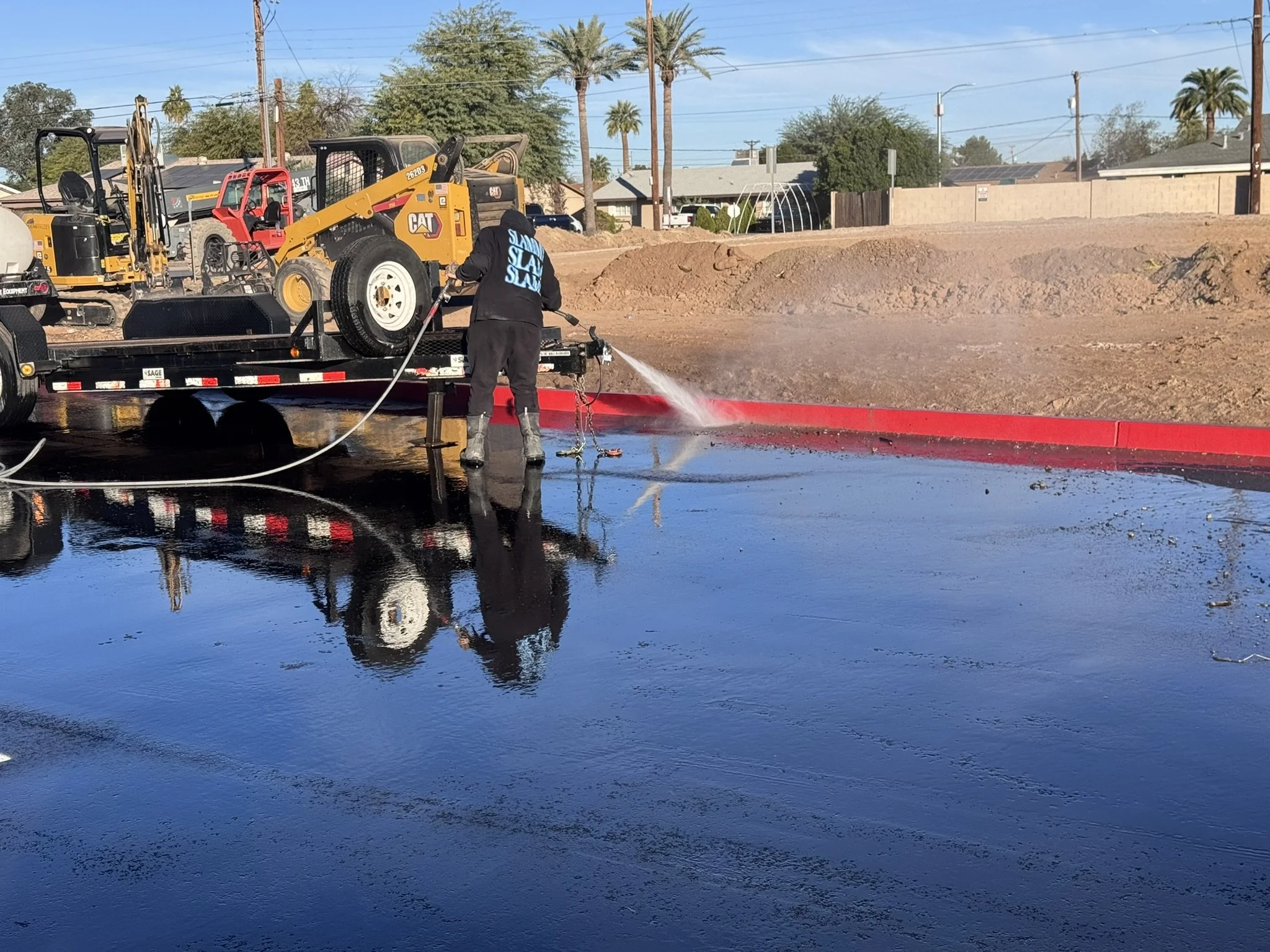 A person spraying water on a puddle near flood barriers, with construction equipment and palm trees in the background.