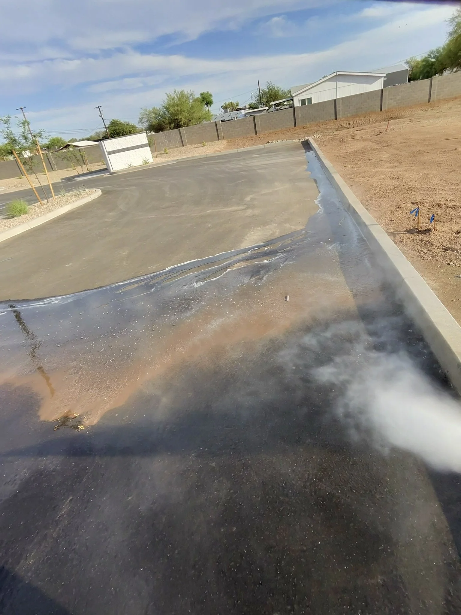 Freshly paved asphalt surface with a water drainage area, small plants, and a sidewalk in a suburban area with a residential fence and houses in the background.