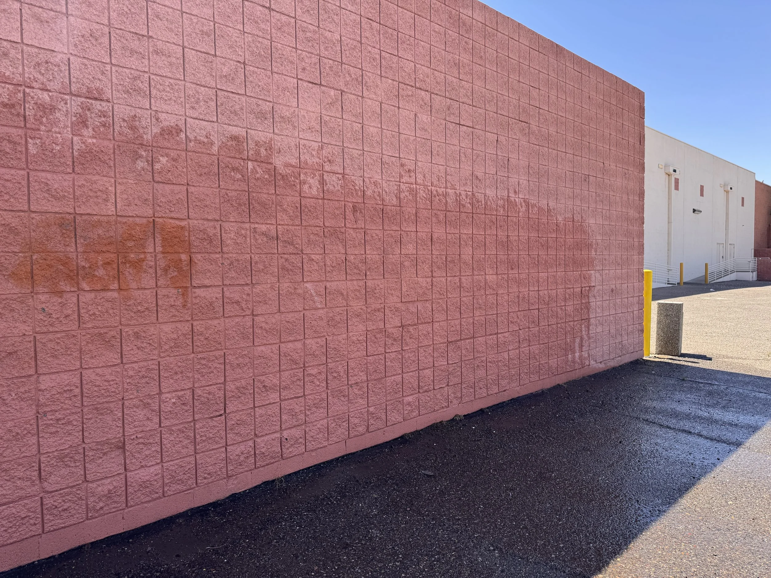 A pink textured concrete wall in an outdoor area with clear blue sky above, part of a building with stairs and yellow safety bollards in the distance.